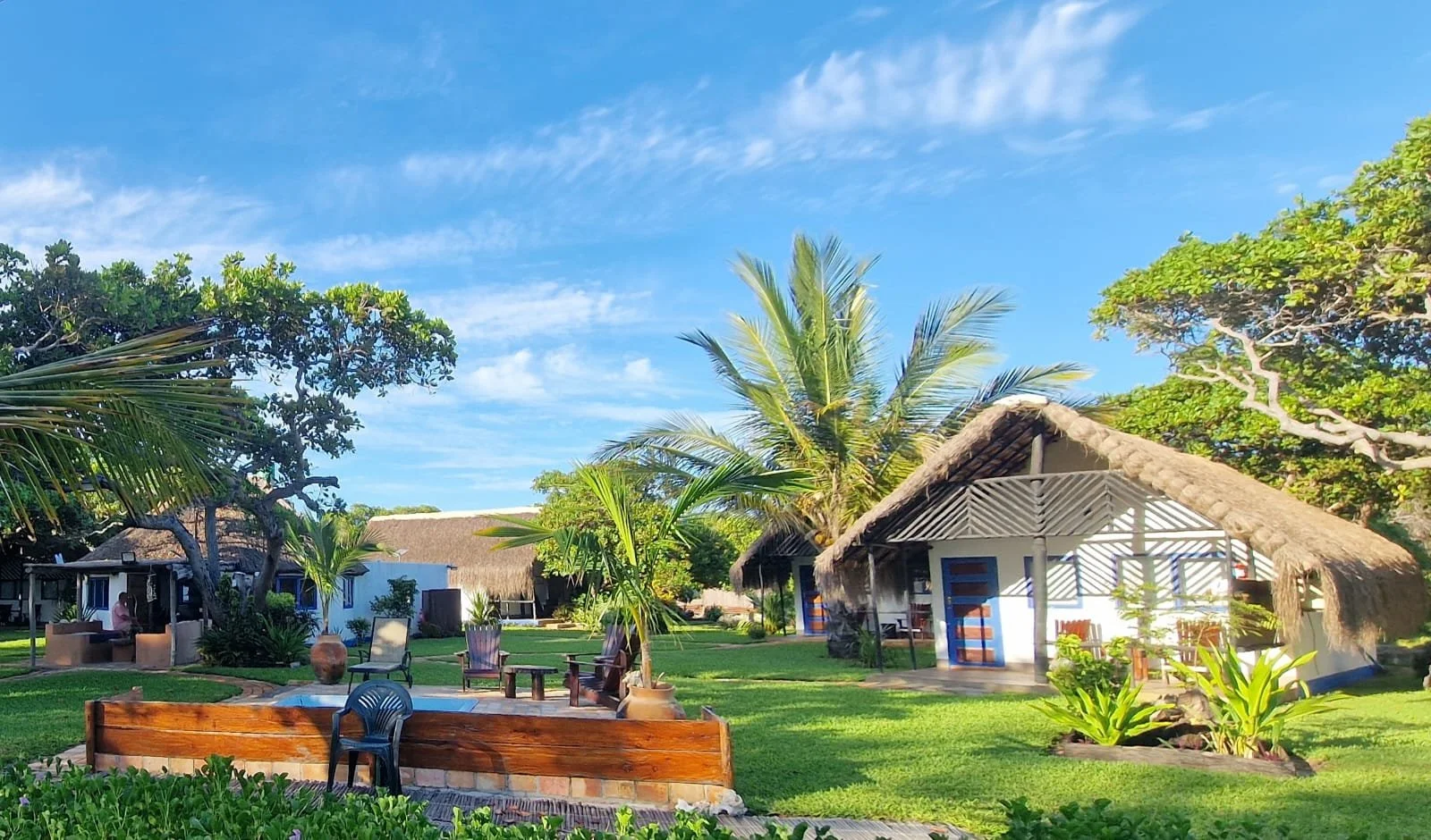 Tropical resort with thatched-roof cabins, lush green grass, and a variety of palm trees under a bright blue sky with scattered clouds in Inhassoro, Mozambique.