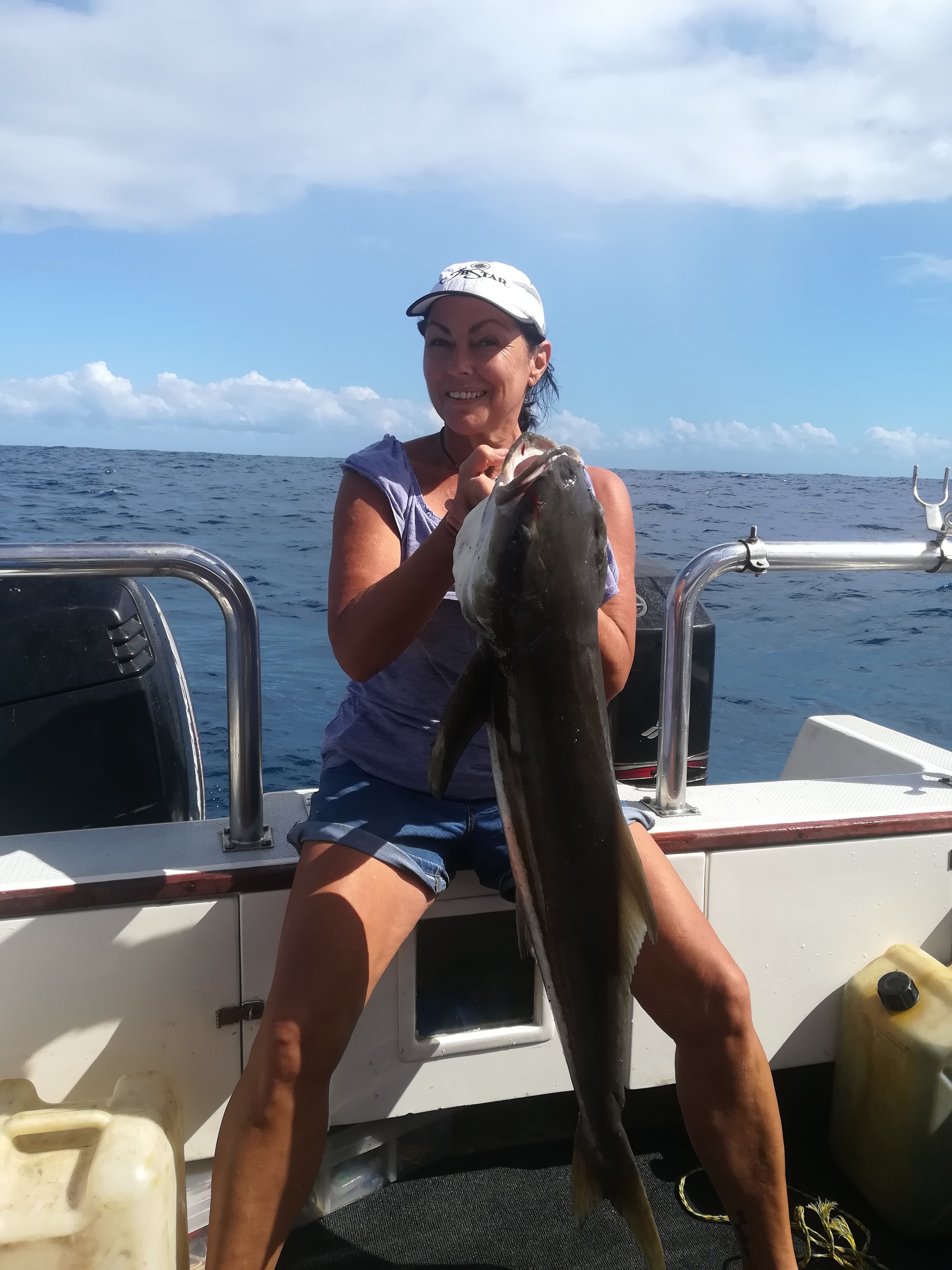 A woman on a boat holding a large fish she caught, with the ocean and partly cloudy sky in the background.
