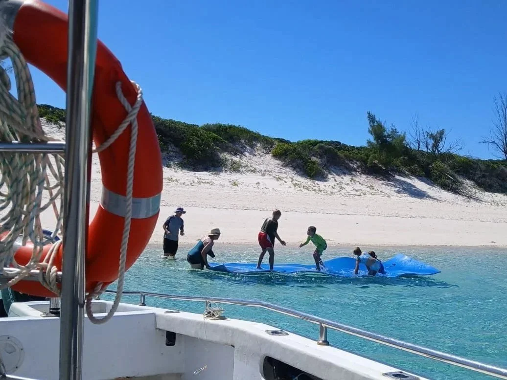 People preparing a blue boat on the beach with a sand dune and trees in the background, seen from a boat with an orange life ring.