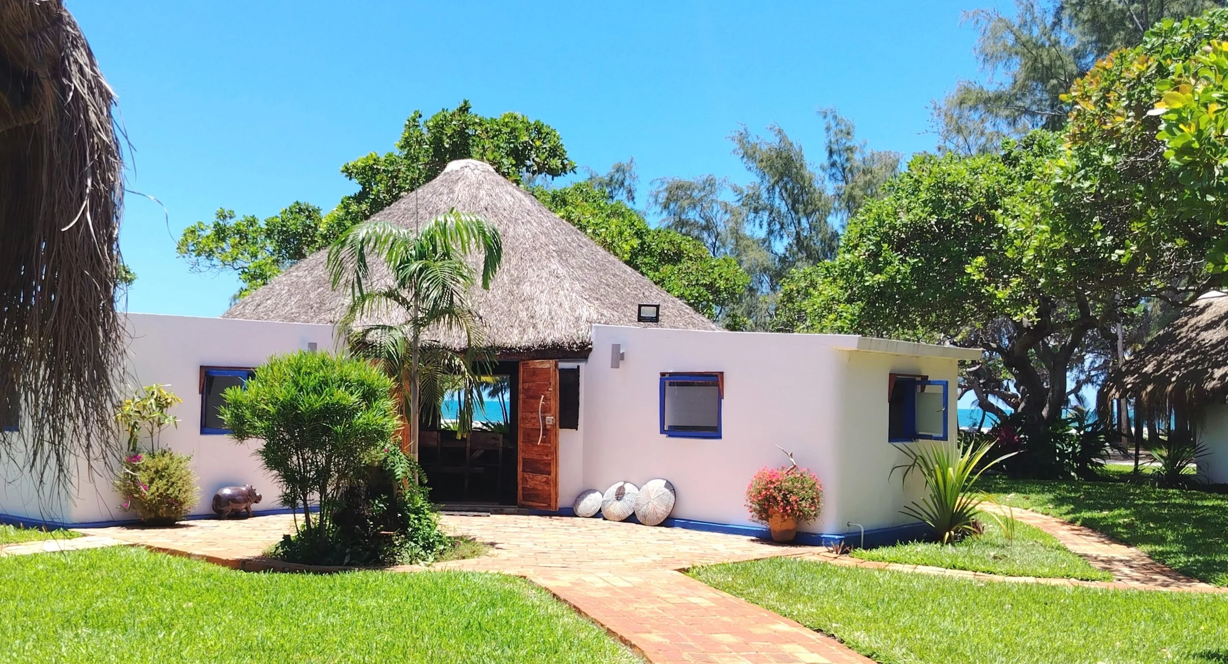 A white building with a thatched roof, surrounded by lush green plants and trees, with a clear blue sky overhead.