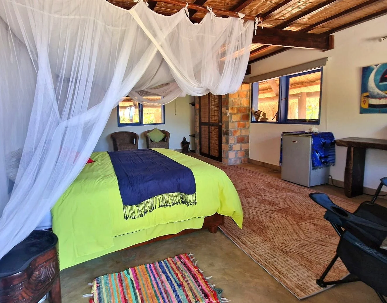 A cozy bedroom with a canopy bed draped with white sheer curtains, a neon green bedspread with a dark blue throw blanket, and wicker chairs near the windows. The room features wooden and brick accents, a small refrigerator, a colorful woven rug, and 