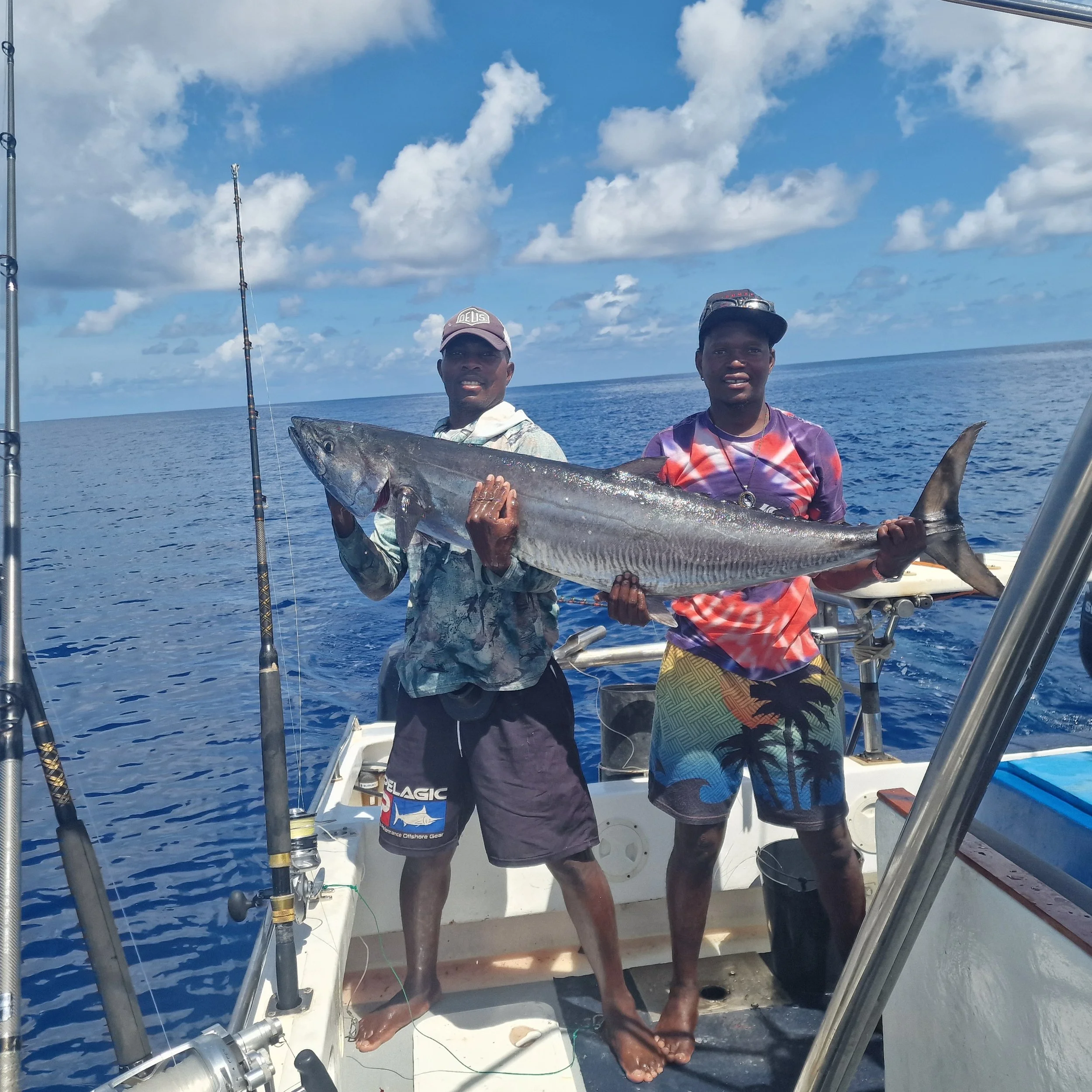 Two young men on a boat holding a large fish, with fishing rods visible, in the open ocean under partly cloudy skies.