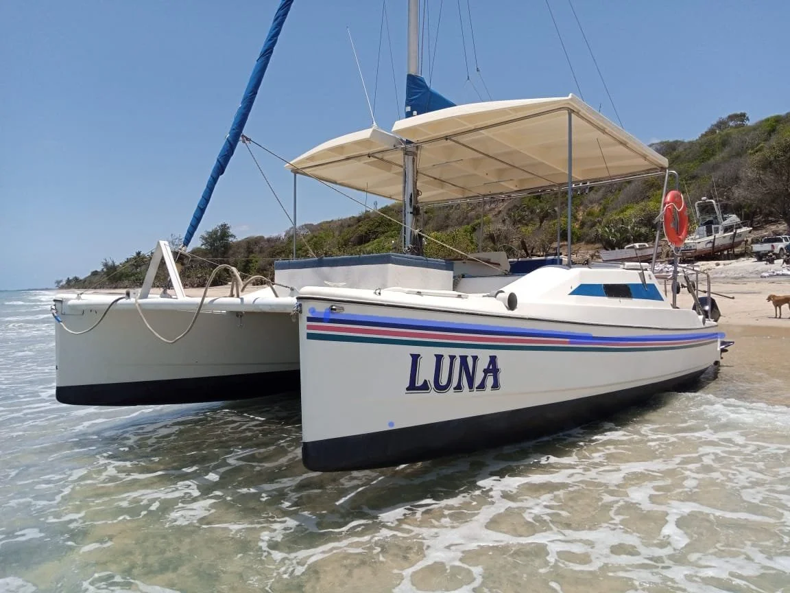 A sailboat with the name Luna on the side, anchored near the shore on a sunny day, with trees and boats in the background in coast of Inhassoro