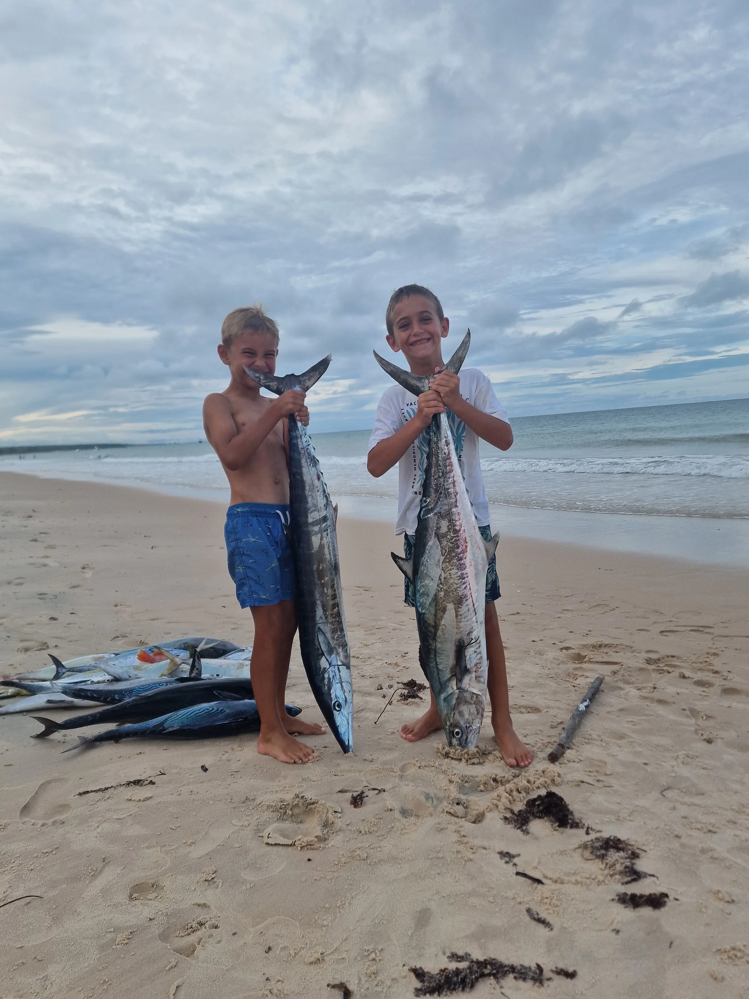 Two young boys on the beach holding large fish they caught, with other fish lying on the sand nearby, and the ocean and cloudy sky in the background.