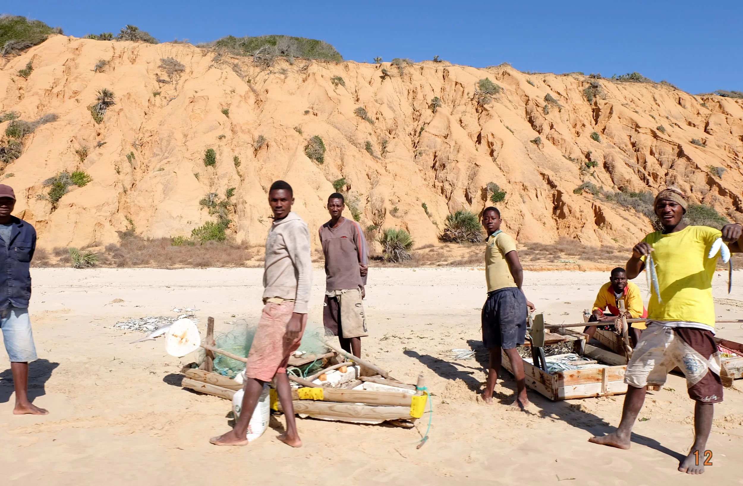 A group of people in a desert landscape with rocky cliffs in the background. Some are standing, some sitting, near wooden structures and debris.