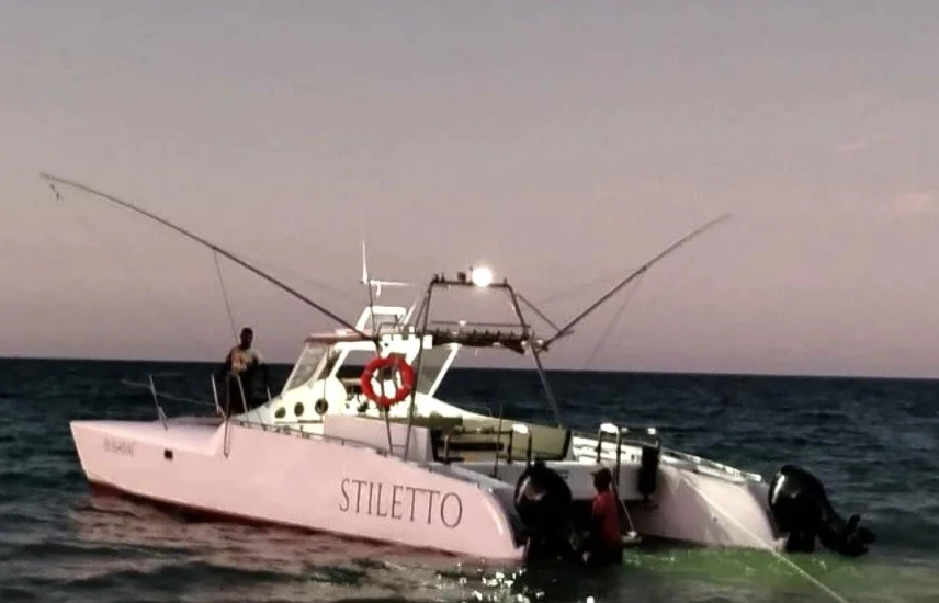A white boat named 'Stiletto' in the ocean at dusk with two people on board, one sitting at the side and one near the motor, against a cloudy sky and open water.
