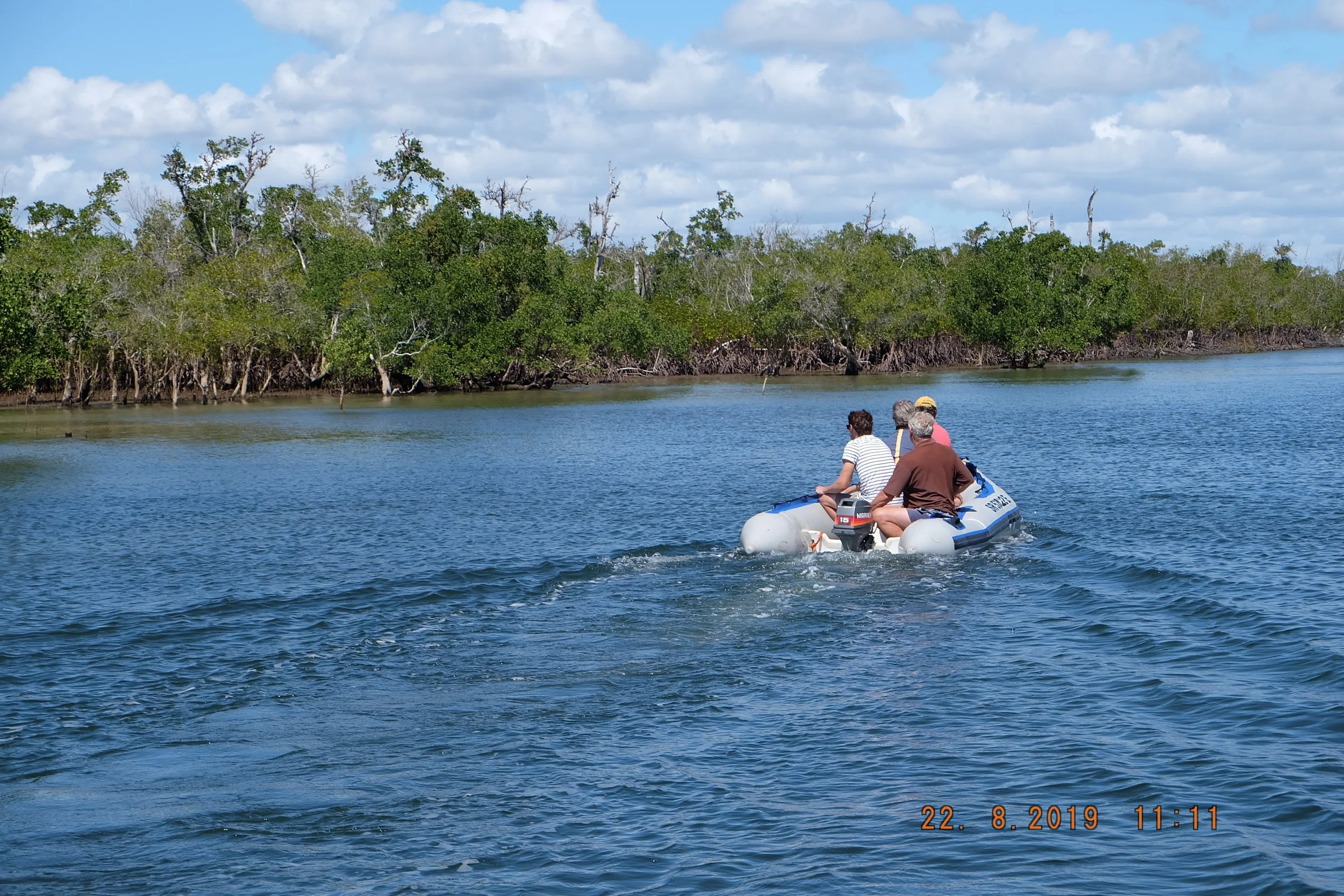 People riding an inflatable boat on a waterway surrounded by lush green mangrove trees under a partly cloudy sky.