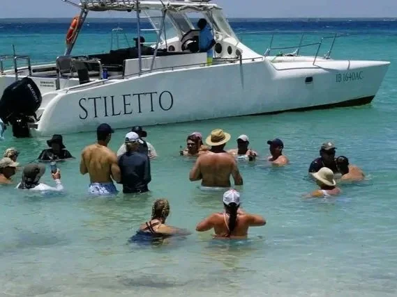 A group of people swimming and standing in shallow turquoise water near a white boat named 'Stiletto' at the beach.