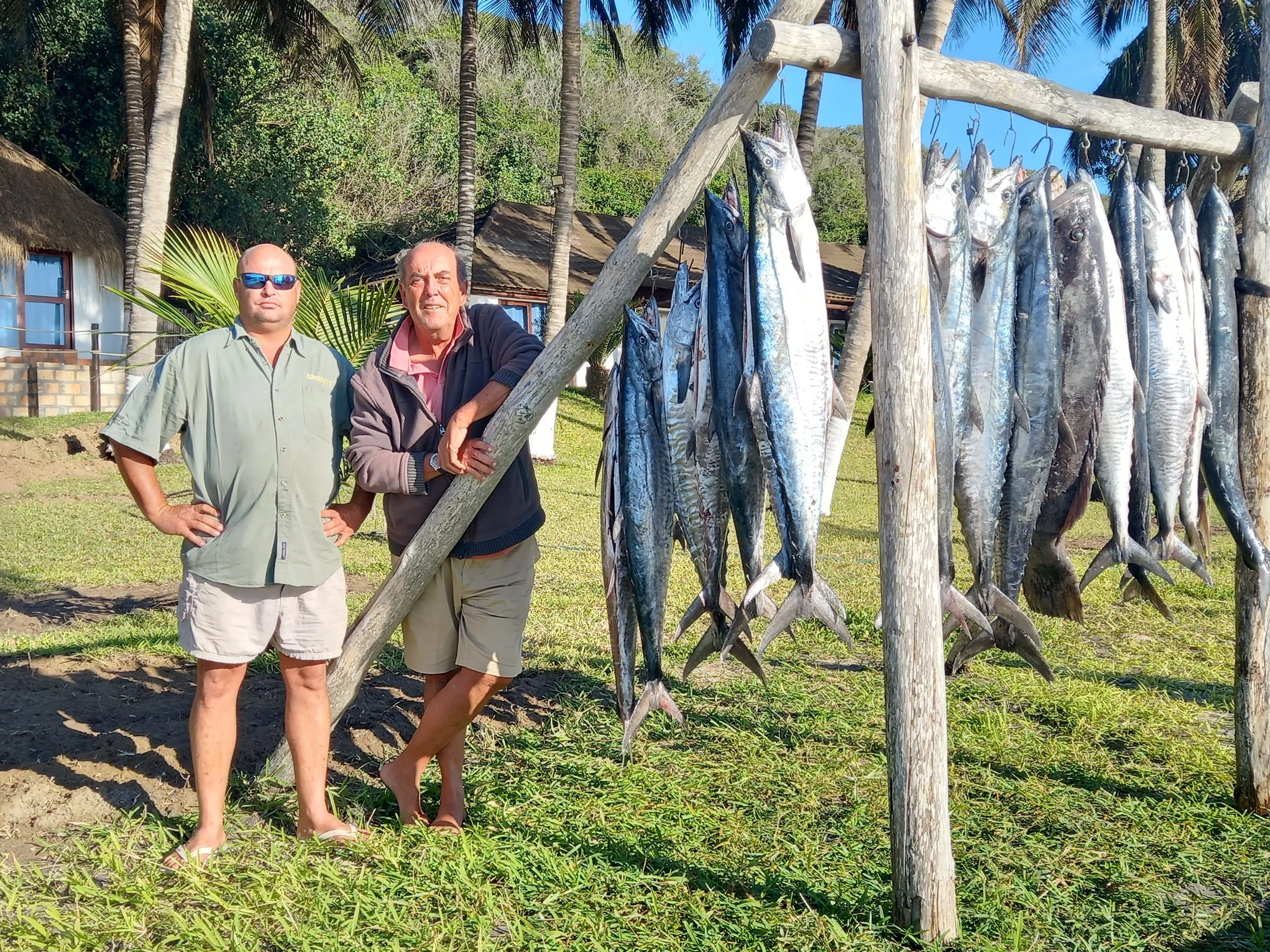 Two men standing outdoors near a wooden structure with dozens of fish hanging on hooks, surrounded by palm trees and houses with thatched roofs.