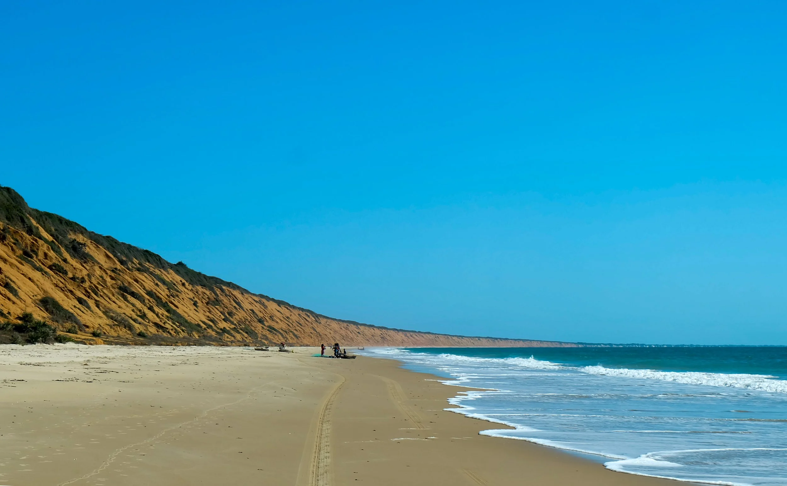 A beach with a sandy shore, some people near the water, and tall, rugged cliffs in the background under a clear blue sky.