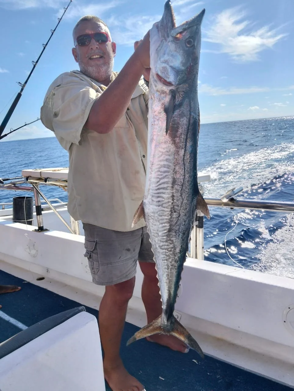 Smiling man in sunglasses holding a large fish on a boat at sea with a fishing rod in the background, under a blue sky with some clouds.