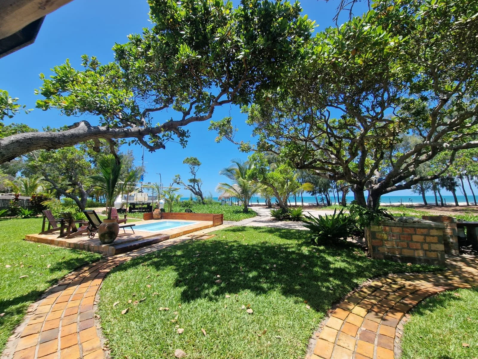 A backyard with a brick pathway, a small pool with lounge chairs, surrounded by green grass, trees, and palm trees, overlooking a beach with the ocean in the background under a clear blue sky in Inhassoro, Mozambique.