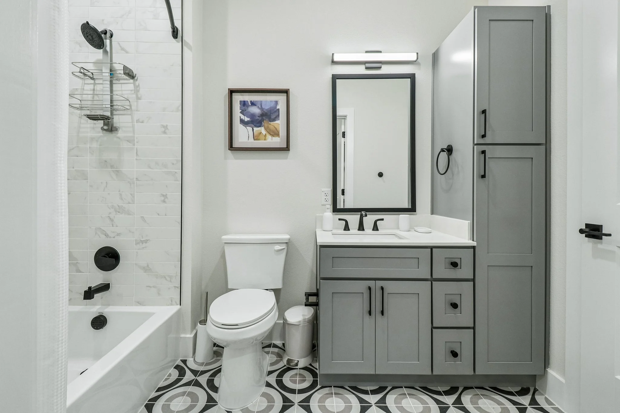 Modern small bathroom with a shower, toilet, and gray vanity with black hardware, white walls, framed artwork, and patterned floor tiles.