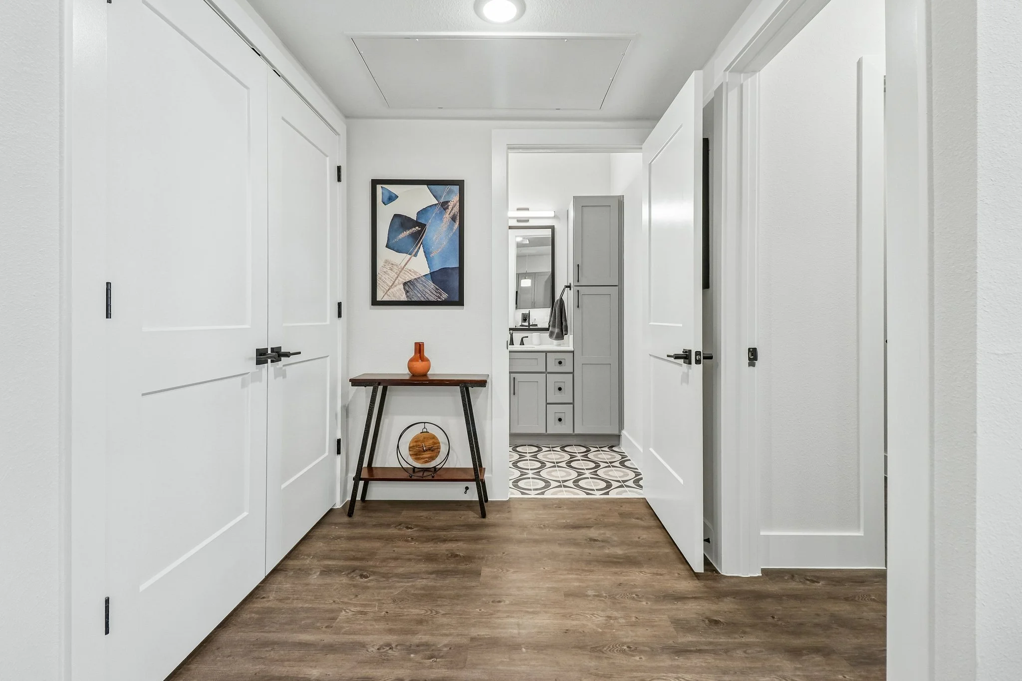 View of a modern, bright hallway with white walls and doors, a wooden floor, a small table with a vase, abstract art painting, and mirror, leading to a bathroom with vanity and towels.