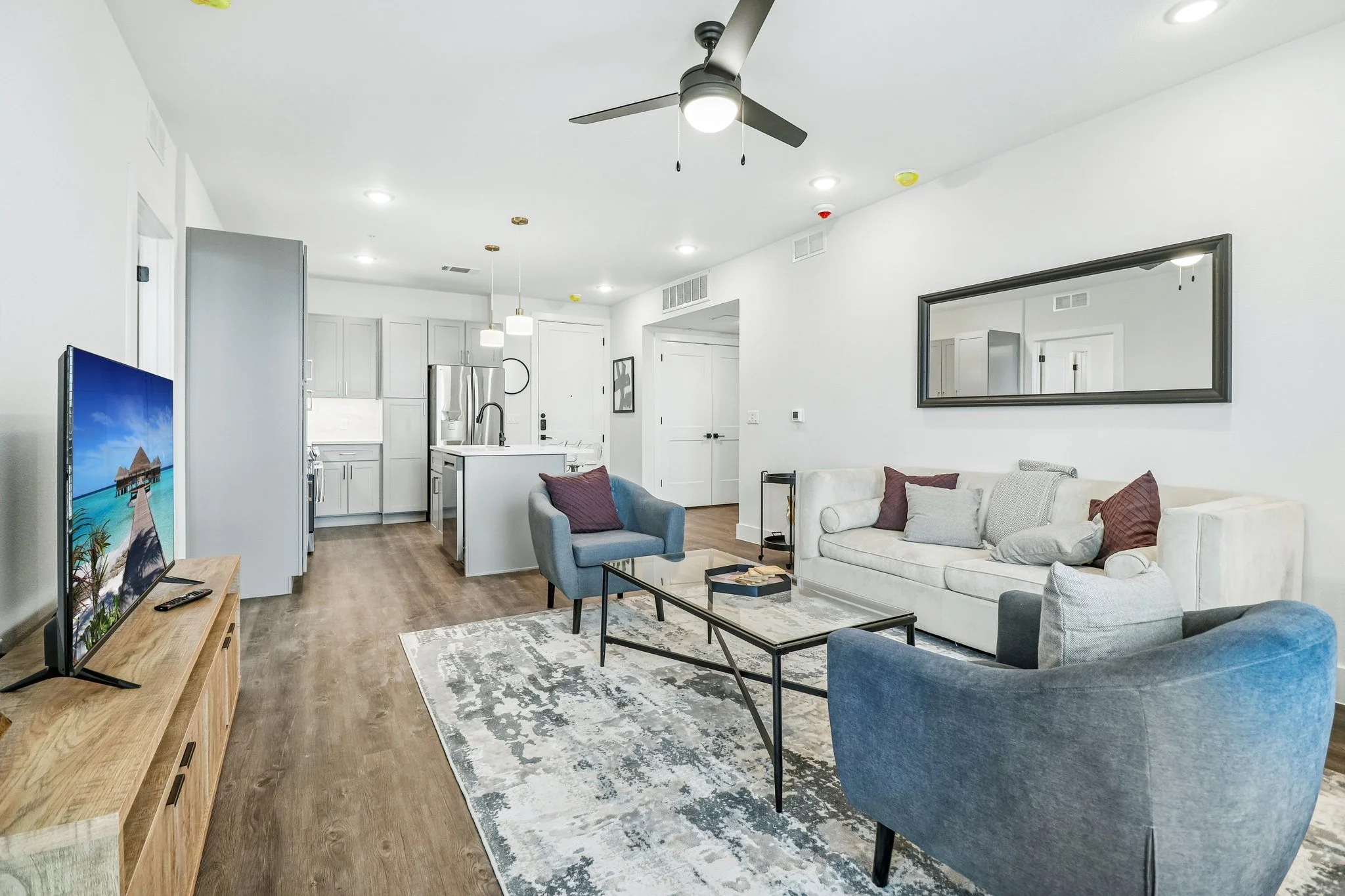 Interior view of a modern living room connected to a kitchen, with white walls, hardwood floors, a large beige sofa with pillows, a blue armchair, a glass coffee table, a wooden TV stand with a flat-screen TV, and a large rectangular mirror on the wa