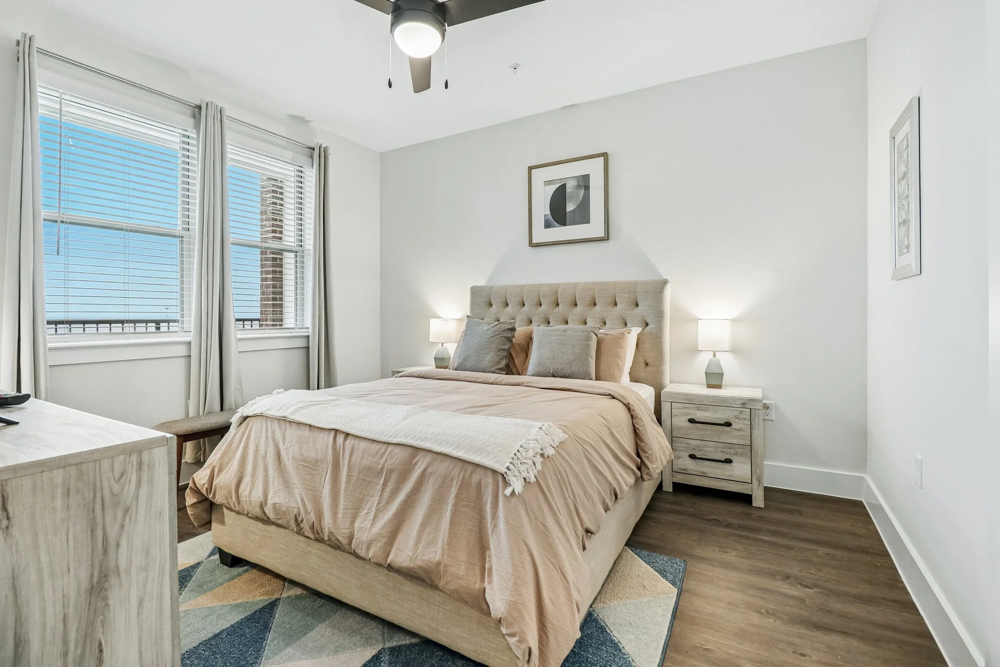 A bedroom with a queen-sized bed with beige bedding and multiple pillows, two nightstands with lamps, a framed abstract art piece on the wall, large windows with white blinds and curtains, a ceiling fan with lights, and a colorful geometric rug.
