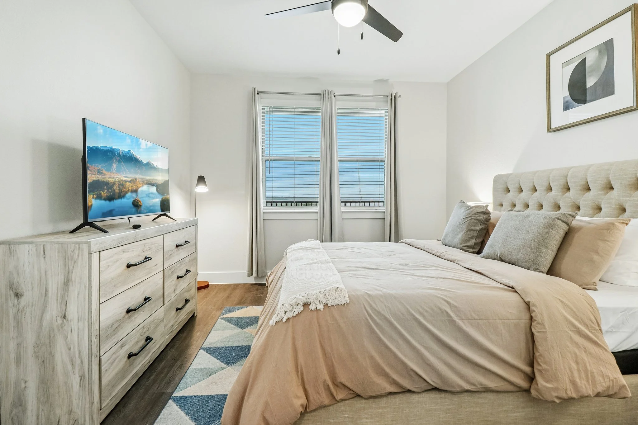 A bedroom with a bed, a dresser with a TV on top, a ceiling fan, and two windows with blinds and curtains.