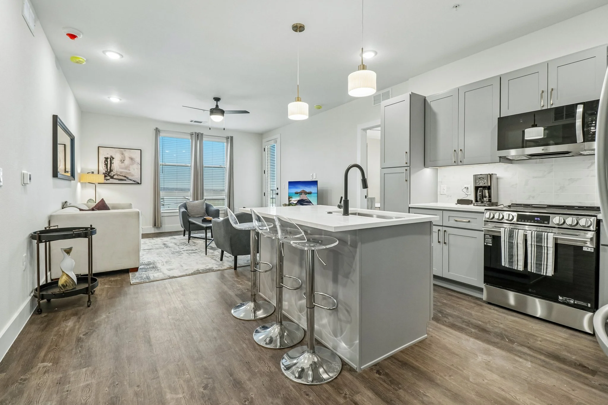 Open-concept kitchen and living room with white cabinets, stainless steel appliances, gray bar stools, a white island, a gray sofa, a wall-mounted TV, windows with blinds, pendant lights, a ceiling fan, and wood flooring.