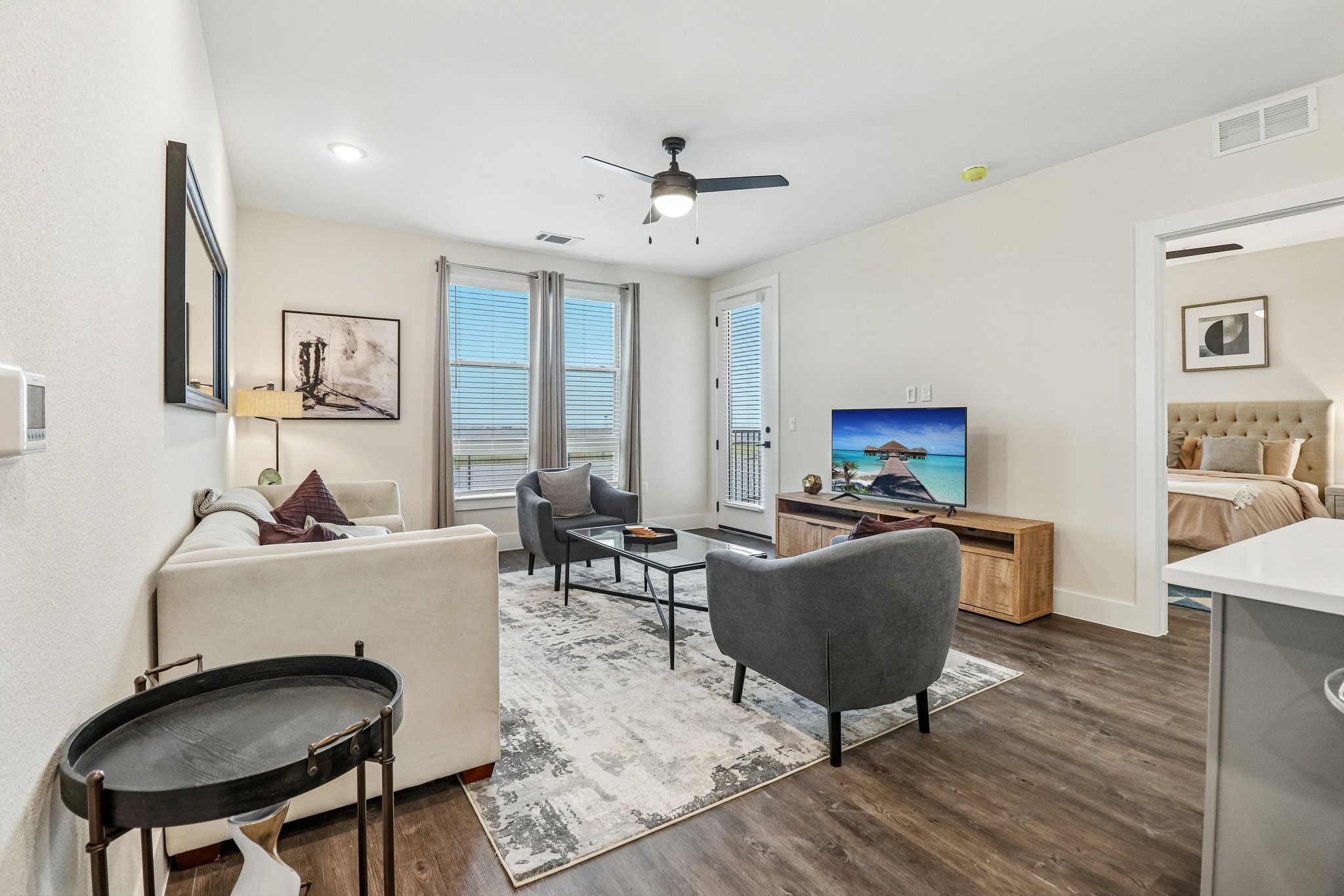 A modern living room with white walls, a ceiling fan, large windows with curtains, and a mix of contemporary furniture including a sofa, armchairs, a TV on a stand showing a beach scene, and wall art. Part of a bedroom with a tufted headboard is visi