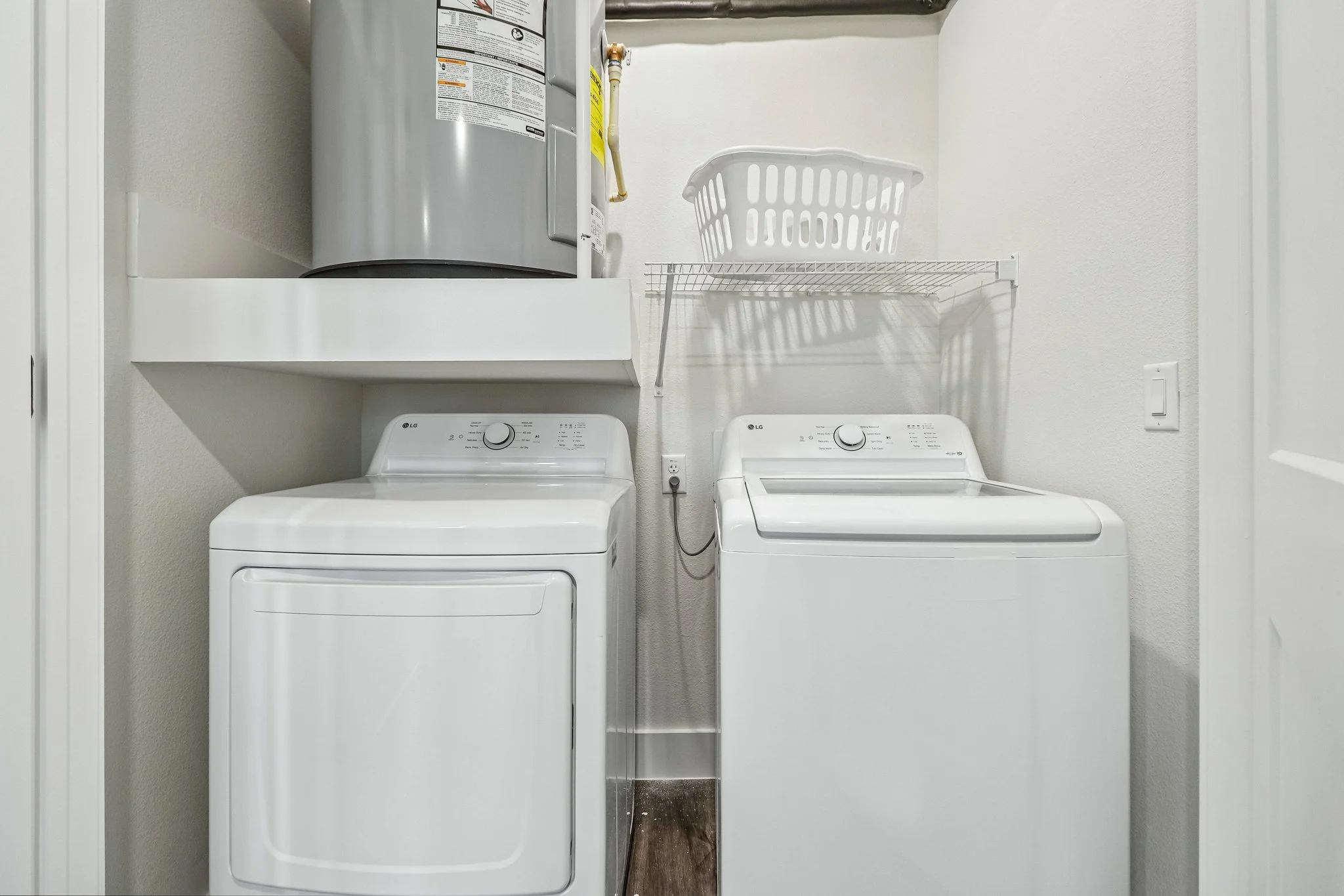 A laundry room with a top-loading washer and dryer, a wire shelf with a laundry basket, and a water heater on a white wall.