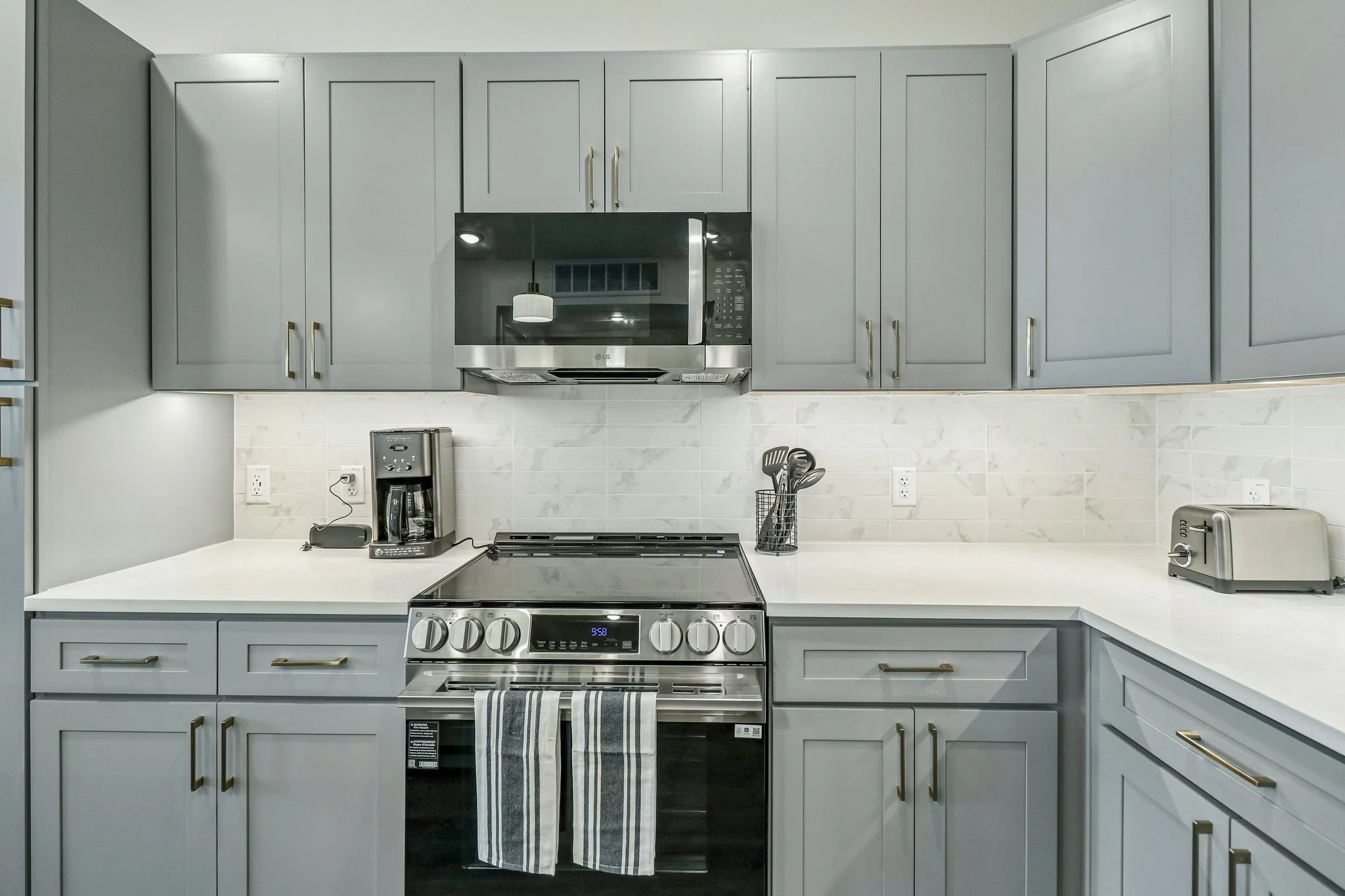 Kitchen with gray cabinets and stainless steel appliances, including a microwave, coffee maker, toaster, and oven, with a white countertop and backsplash.