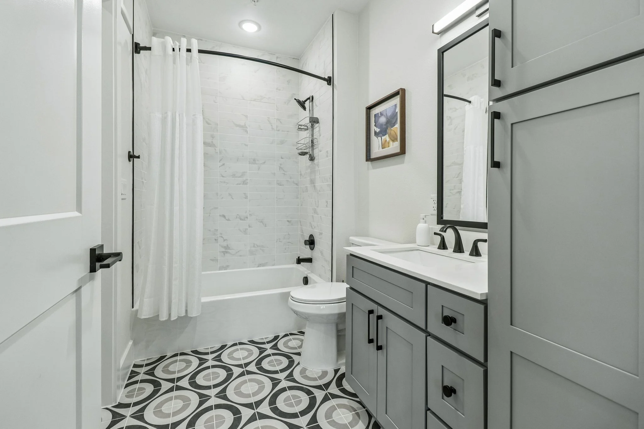 A modern bathroom with a walk-in shower, white marble tiles, a white toilet, gray vanity with black handles, a mirror, and patterned black and white floor tiles.