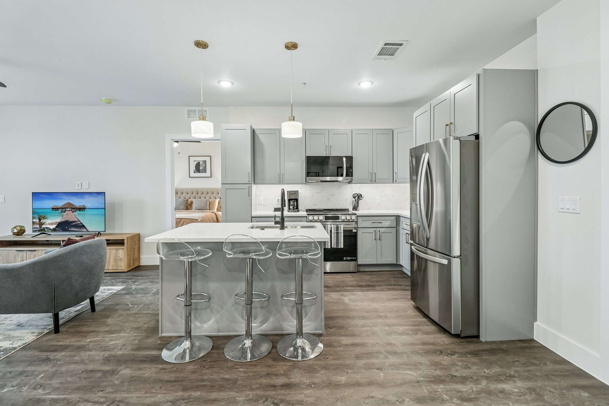 Modern kitchen with gray cabinets, stainless steel appliances, white island with three clear bar stools, and a view into a bedroom with a beige headboard.