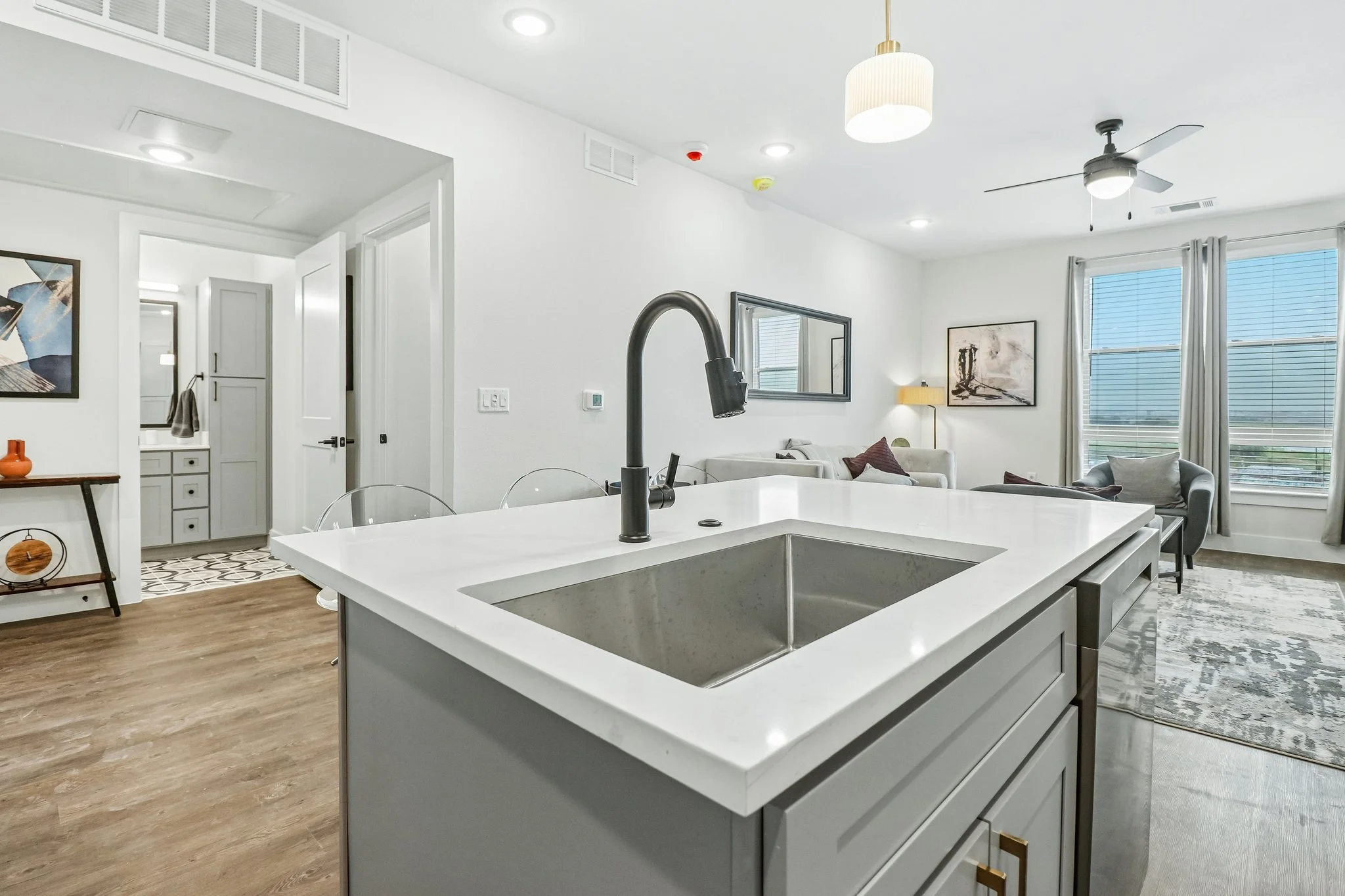 Modern open-concept living space and kitchen in a home with white walls, large windows, and contemporary furniture, including a white kitchen island with a black faucet.
