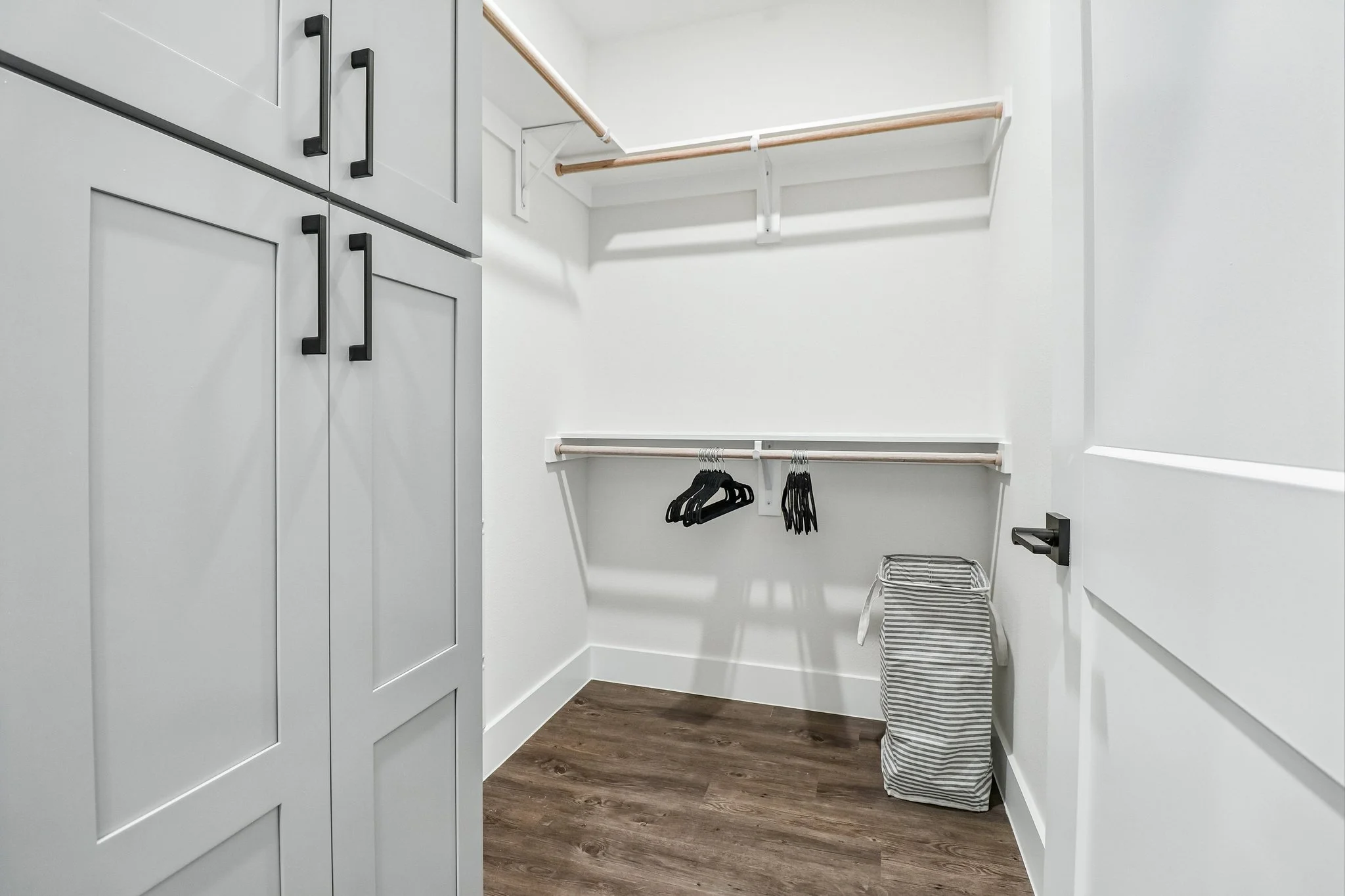 A walk-in closet with white walls, brown wood flooring, white cabinetry with black handles, and wooden hanging rods. There are black hangers hanging from the rods and a striped laundry basket on the floor. A small wire basket is in the corner.