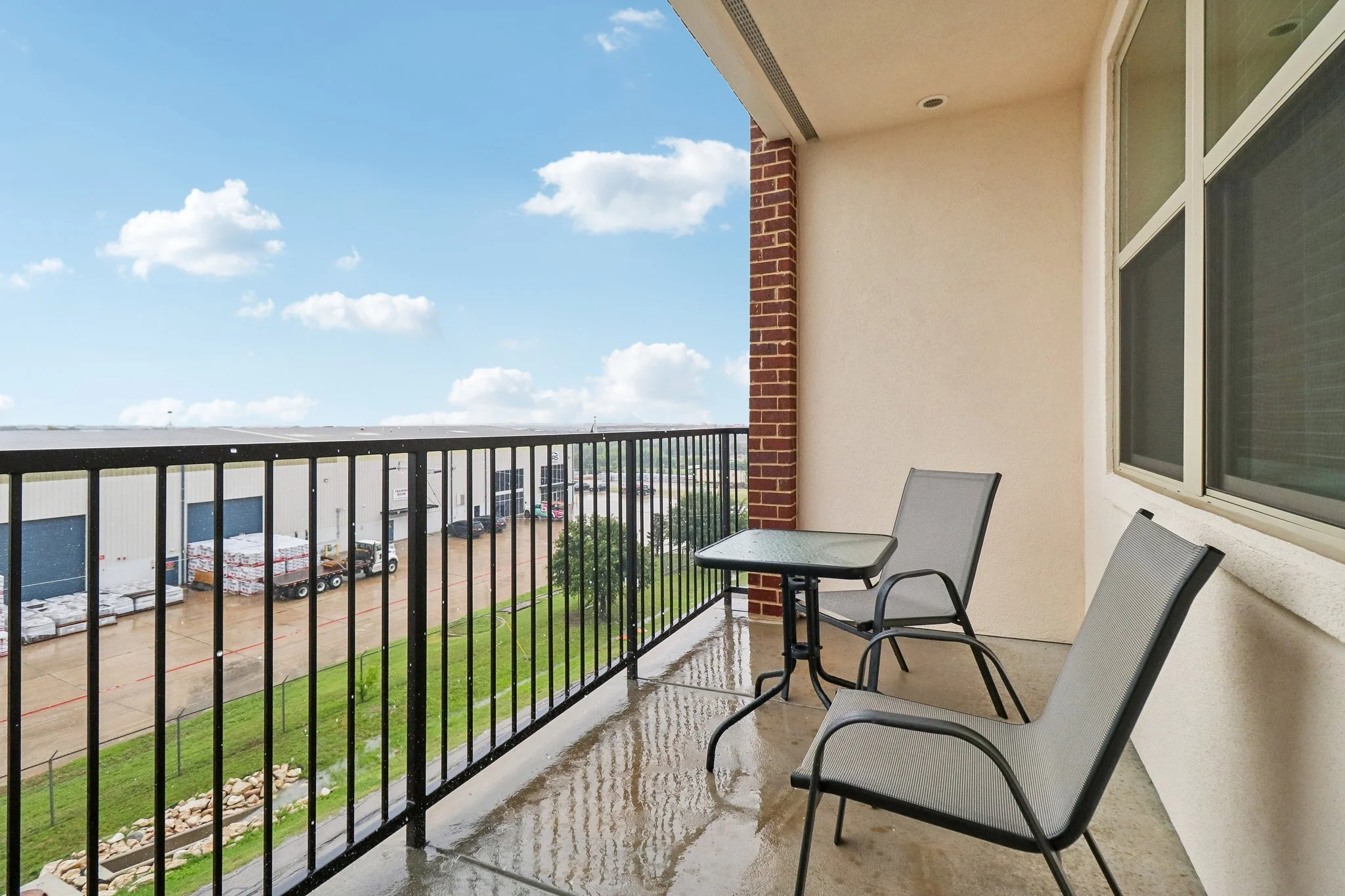 A balcony with wet flooring, two metal chairs and a table, overlooking a parking lot with trucks and buildings under a partly cloudy sky.