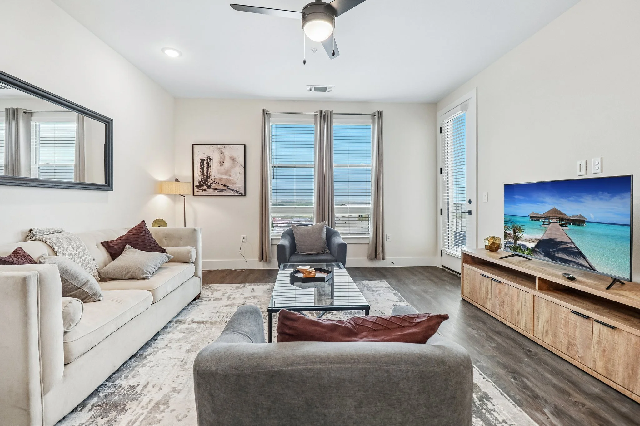 Living room with white sofa, gray armchair, and wooden TV stand with a large flat-screen TV displaying a beach scene. The room features hardwood floors, a ceiling fan, and large windows with blinds and curtains, allowing natural light.