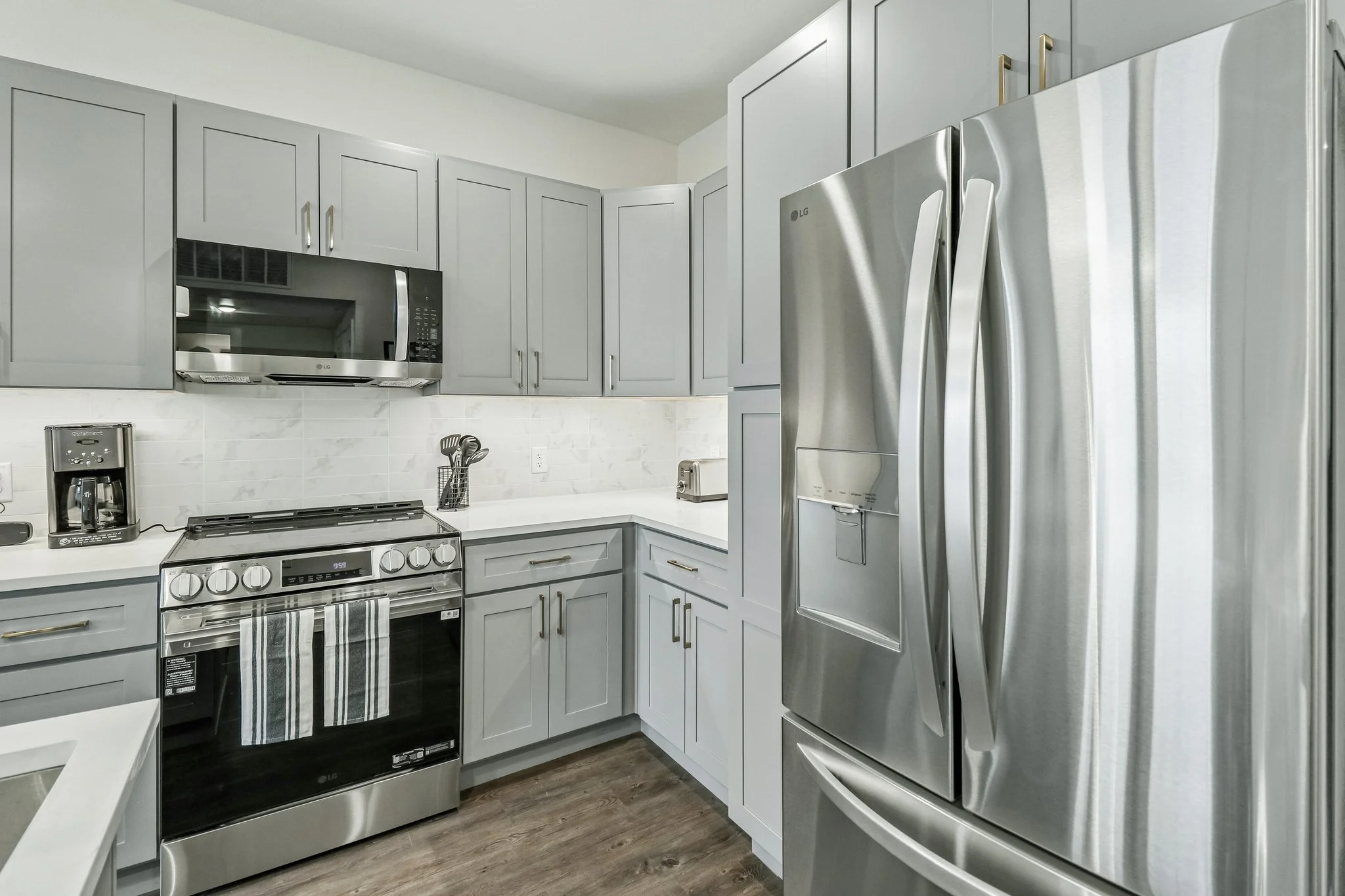 A modern kitchen with gray cabinets, stainless steel appliances including a refrigerator, microwave, and oven, and a white countertop with kitchen utensils and a coffee maker.