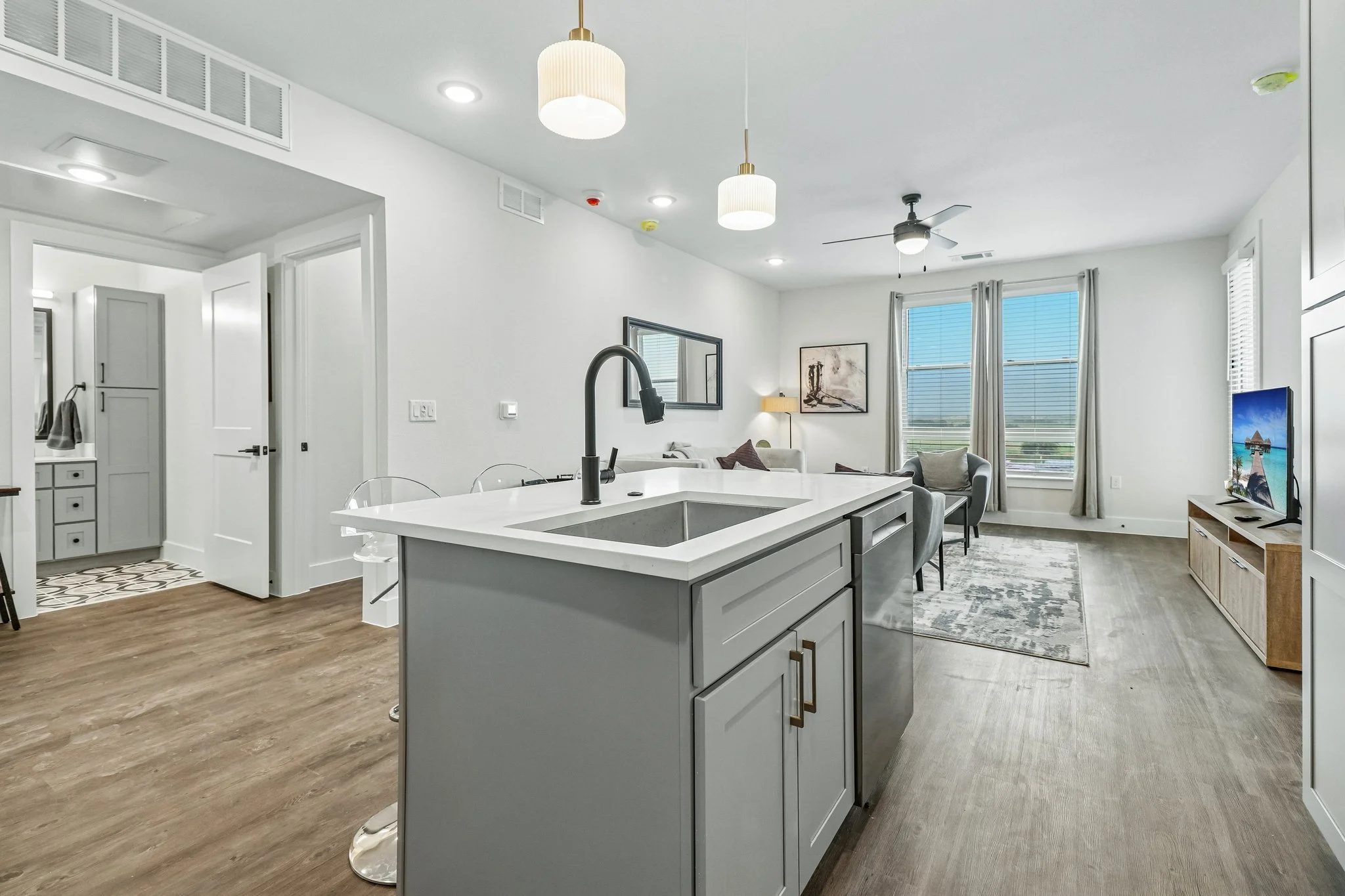 Bright open-concept living space with white walls, large windows with curtains, modern furniture, a kitchen island with a black sink, and a television in the corner.