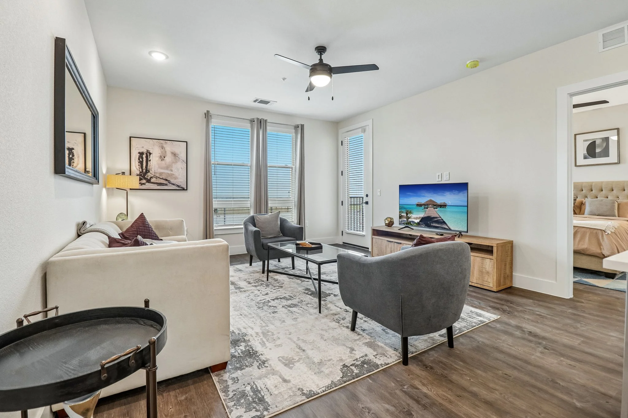 Living room with a beige sofa, gray armchair, wooden TV stand, TV showing a beach scene, framed art on the walls, ceiling fan, sunlight through windows, doorway to a bedroom.