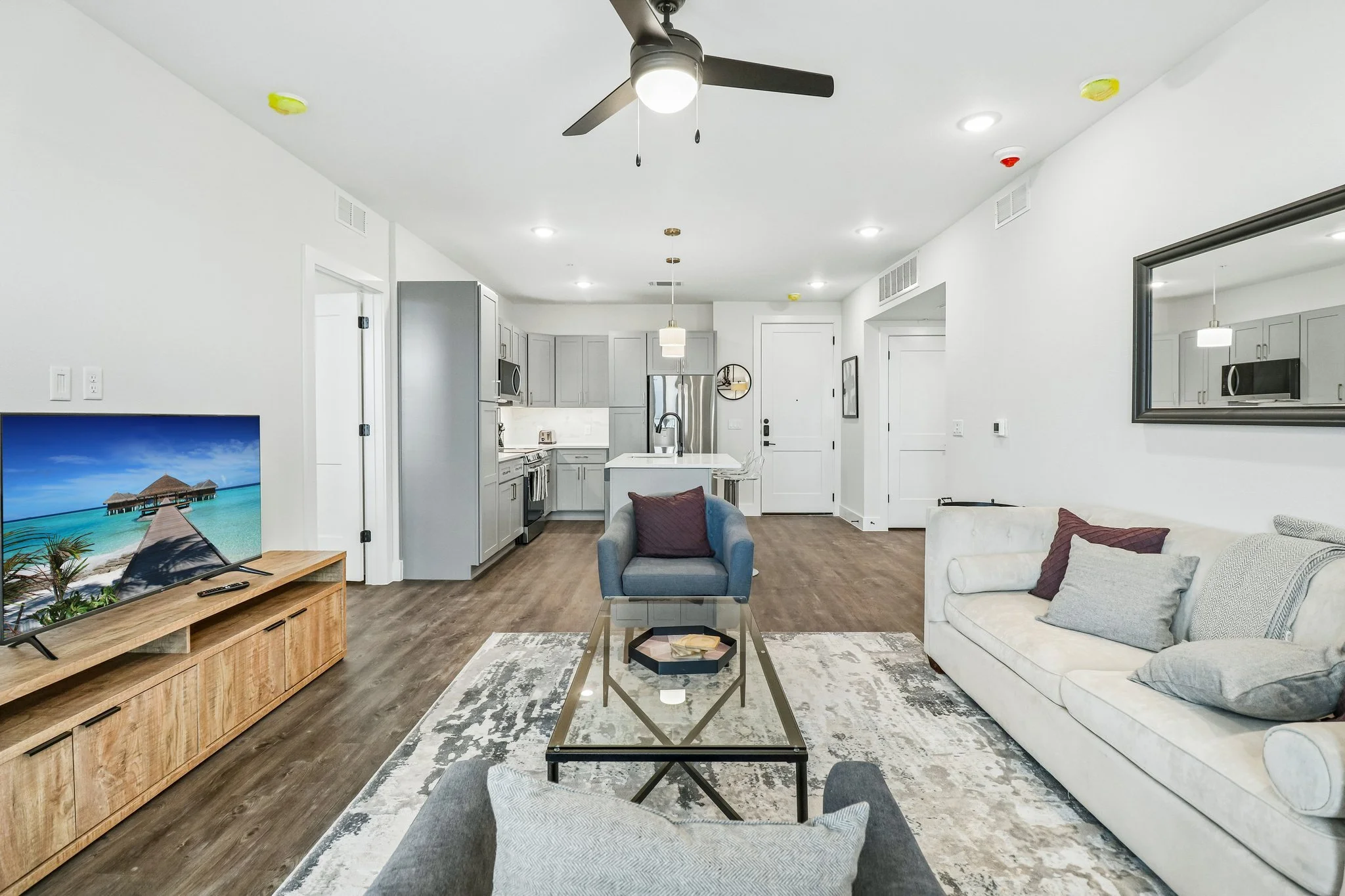 Open-concept living room and kitchen with white walls, gray cabinets, stainless steel appliances, and hardwood flooring. A white sofa with pillows, a blue armchair, and a wooden media console with a television are visible.