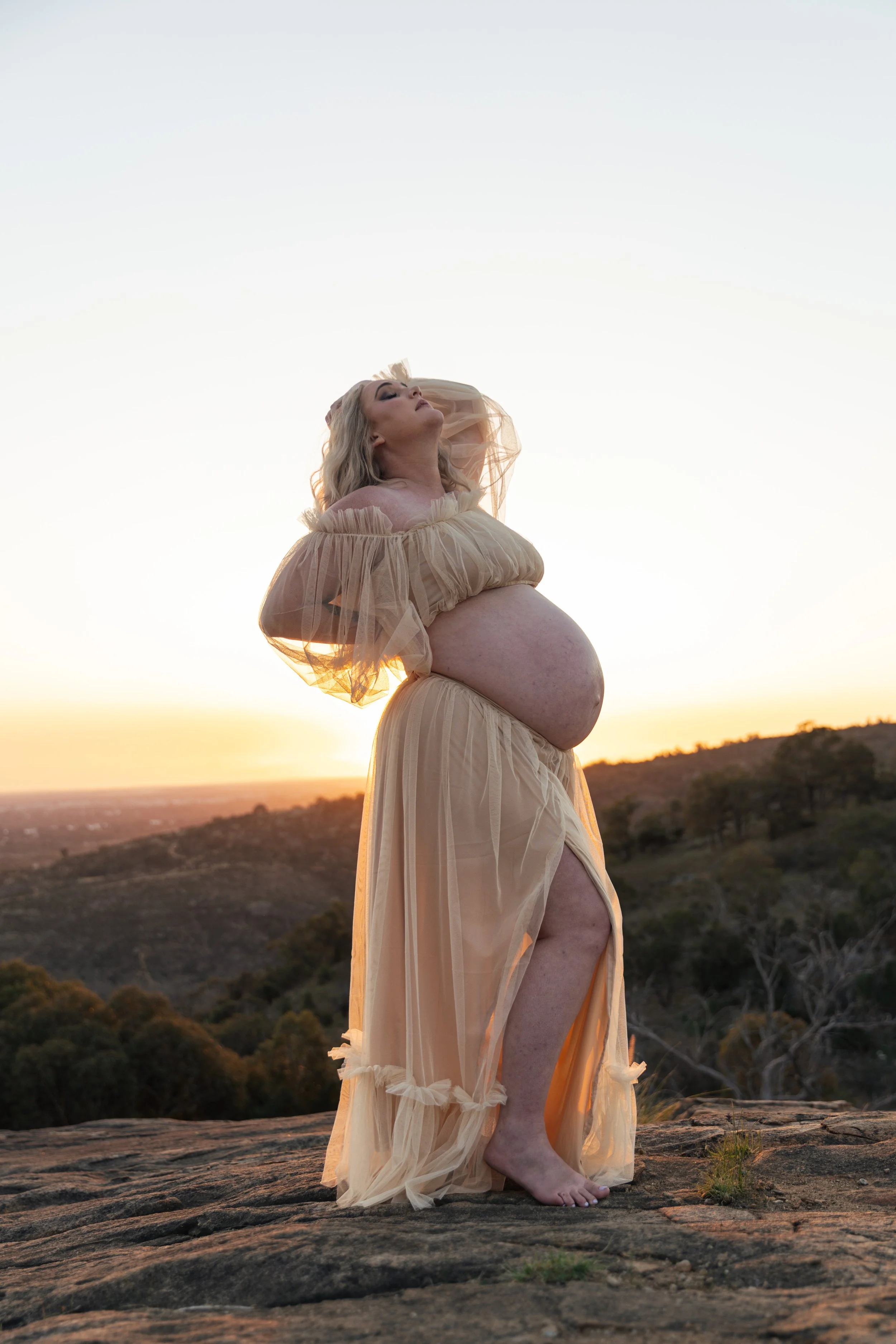 Pregnant woman standing on rocky terrain during sunset, wearing a beige flowing dress with sheer puffed sleeves, with her head tilted back and eyes closed.