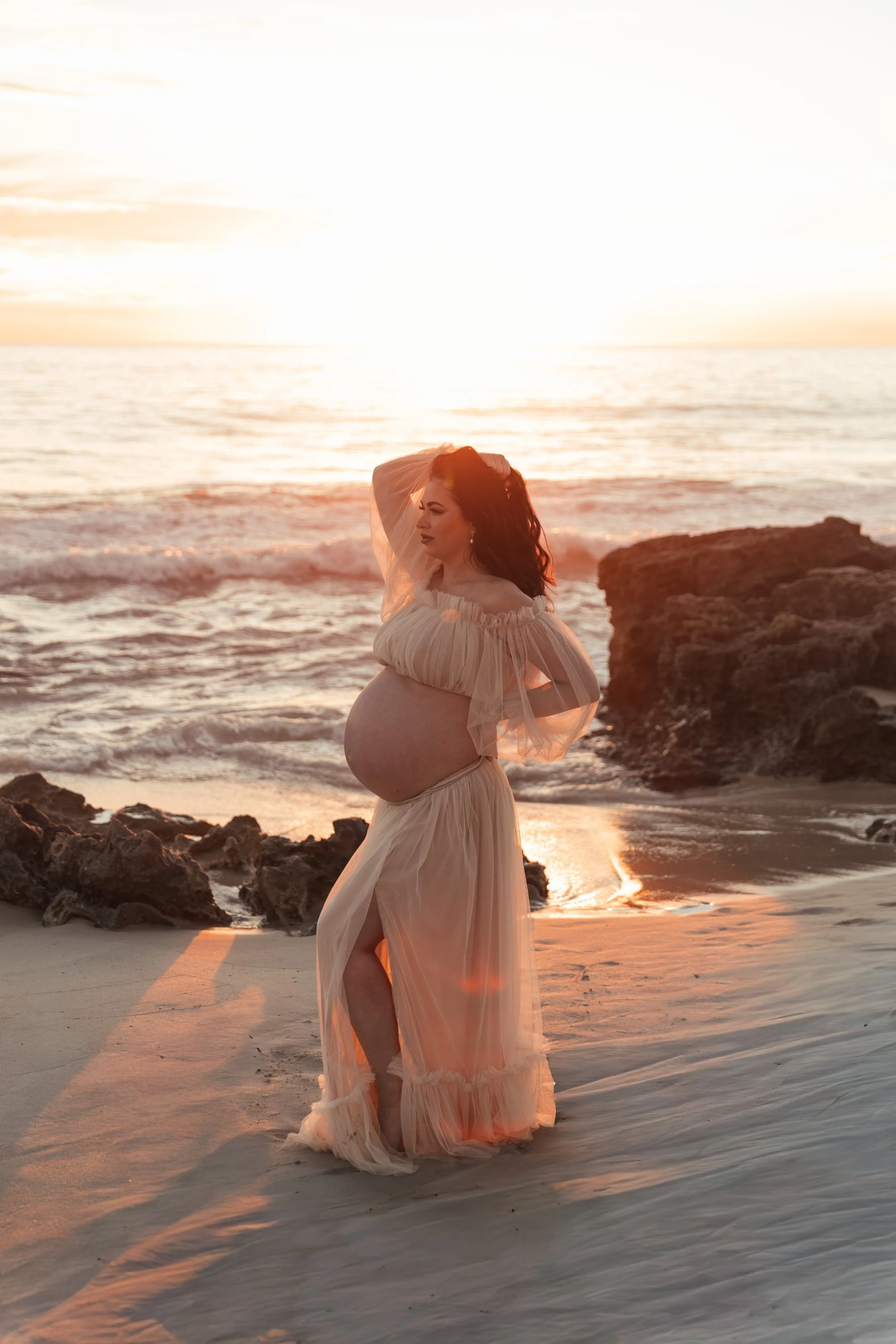 A pregnant woman in a flowing, off-shoulder dress standing on a beach at sunset with ocean waves and rocks in the background.
