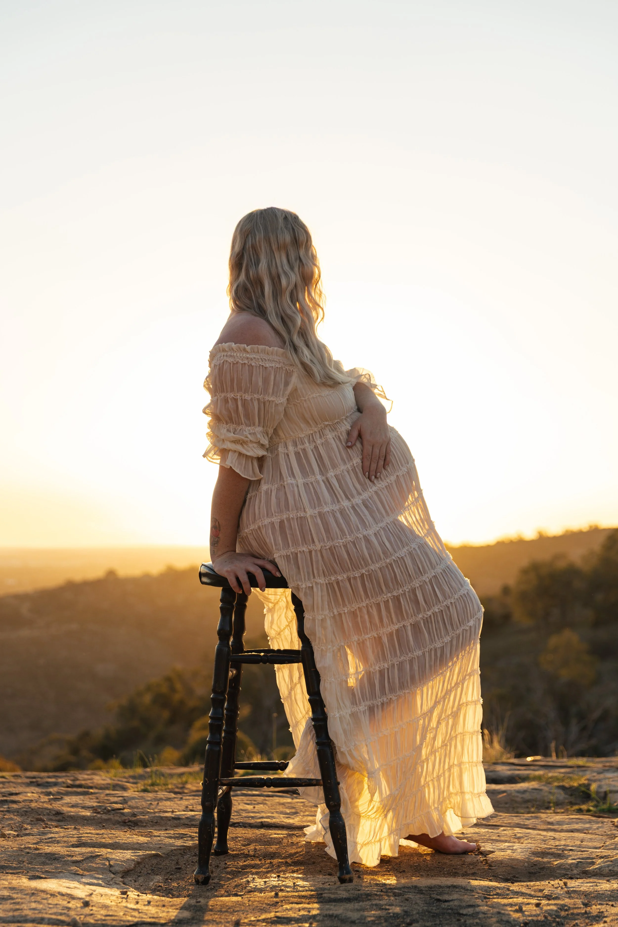 A woman in a flowing beige dress stands outdoors during sunset, holding onto a black wooden chair with one hand and resting her other hand on her pregnant belly. The landscape features rolling hills and a clear sky.