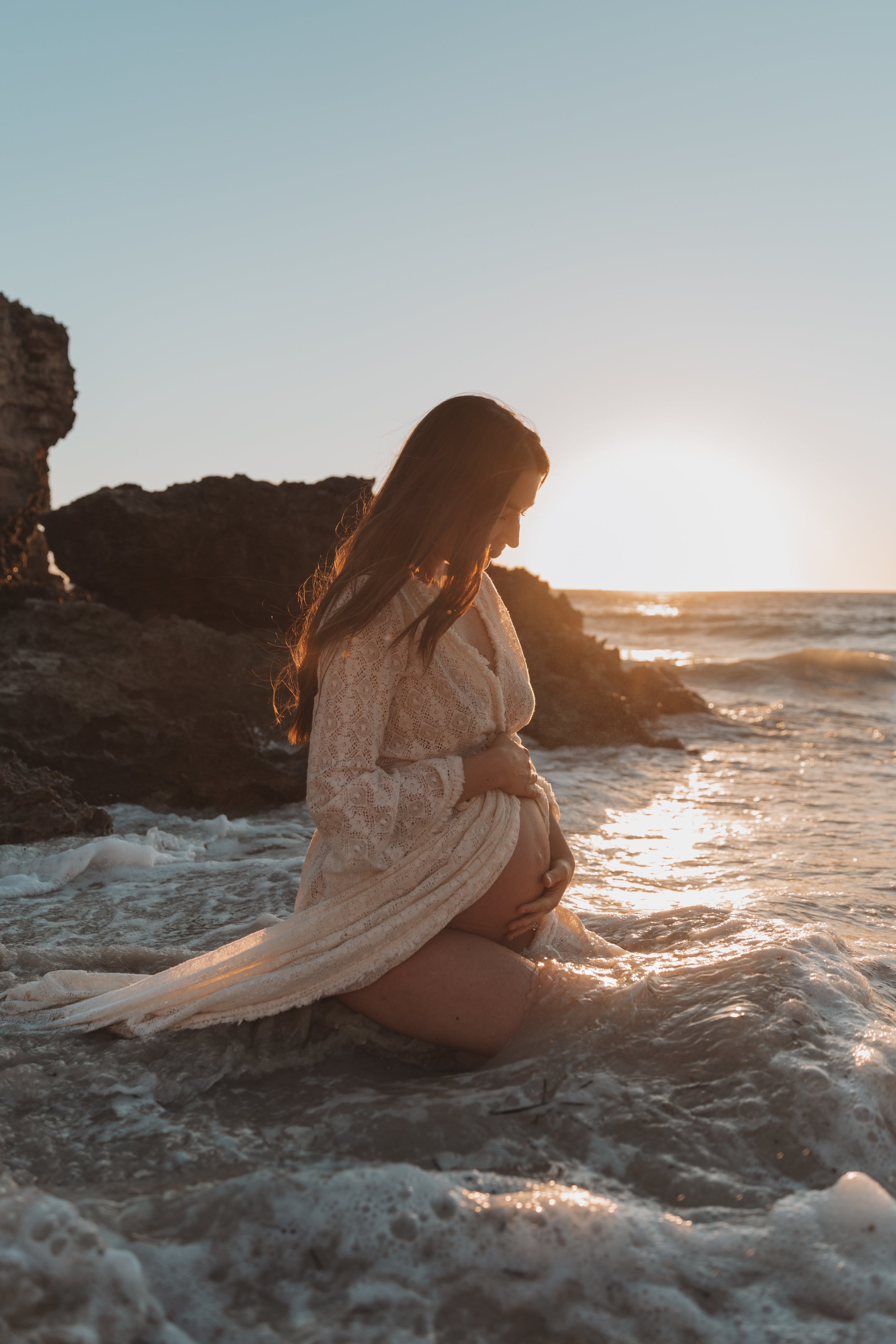 A pregnant woman sitting on a rocky beach at sunset, gently holding her belly and looking down.