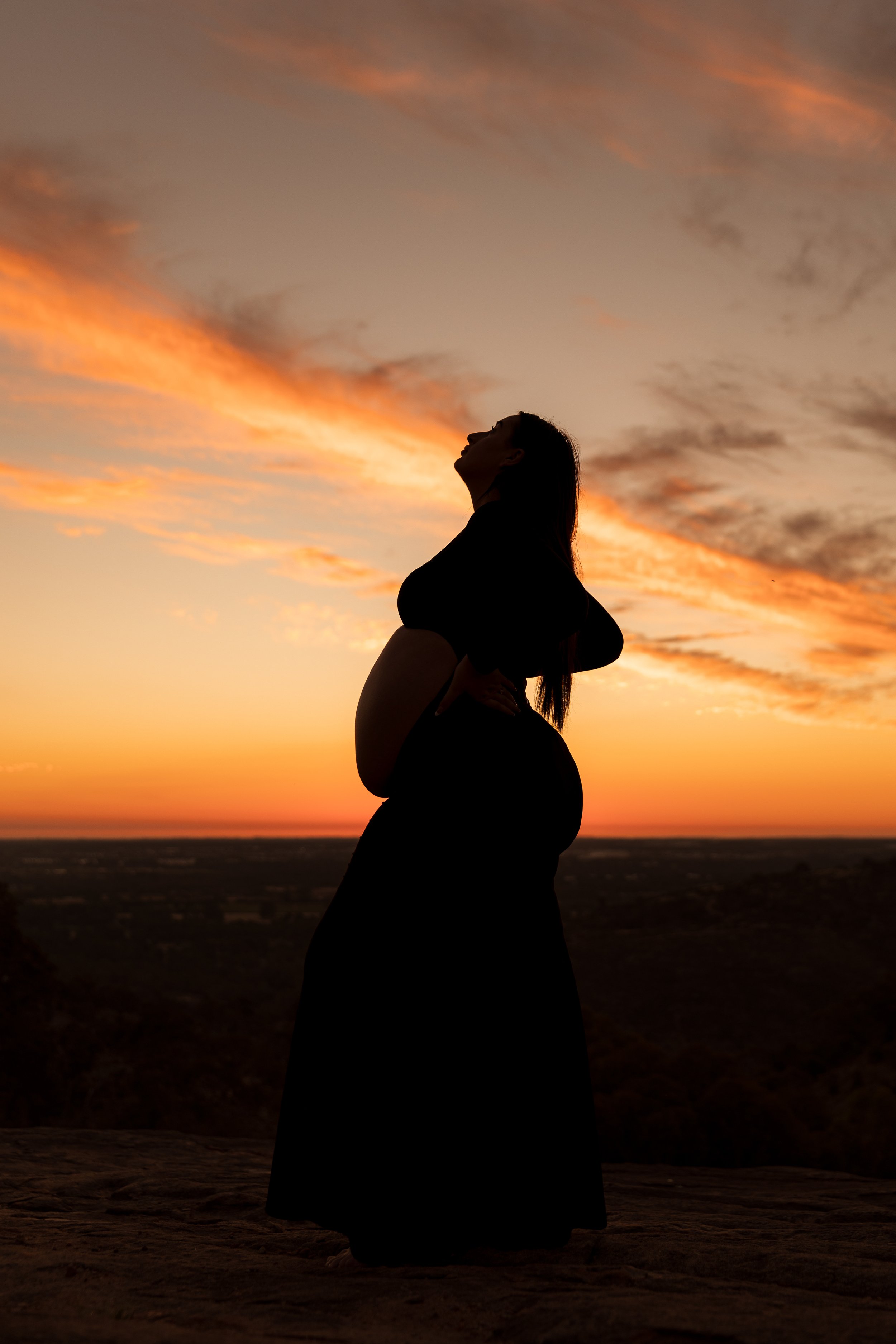 Silhouette of a woman standing with her hand on her stomach, looking up, during a sunset sky with orange and pink clouds.
