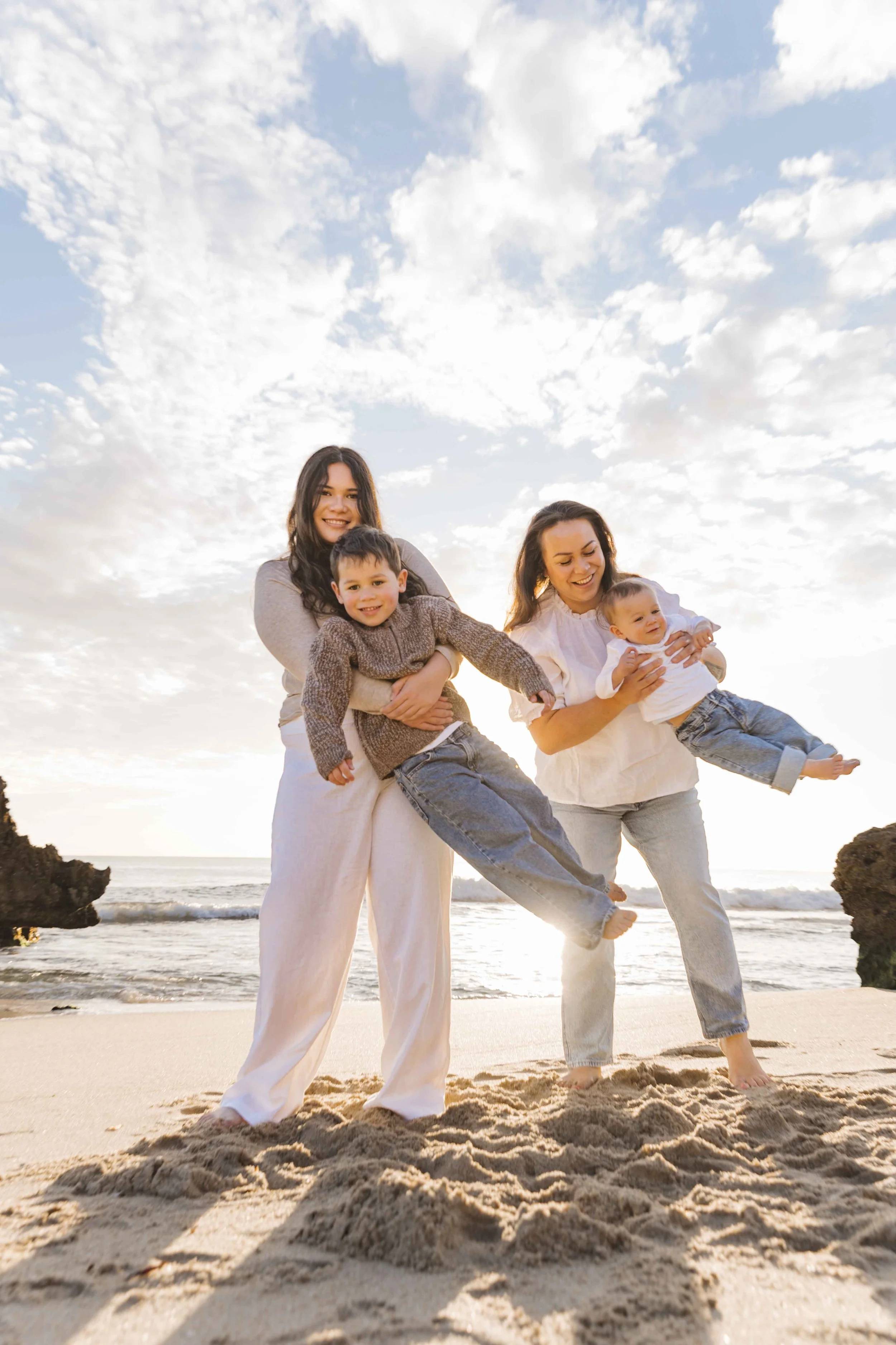 Two women and two children on a beach, holding and playing with each other, with ocean and sky in the background.