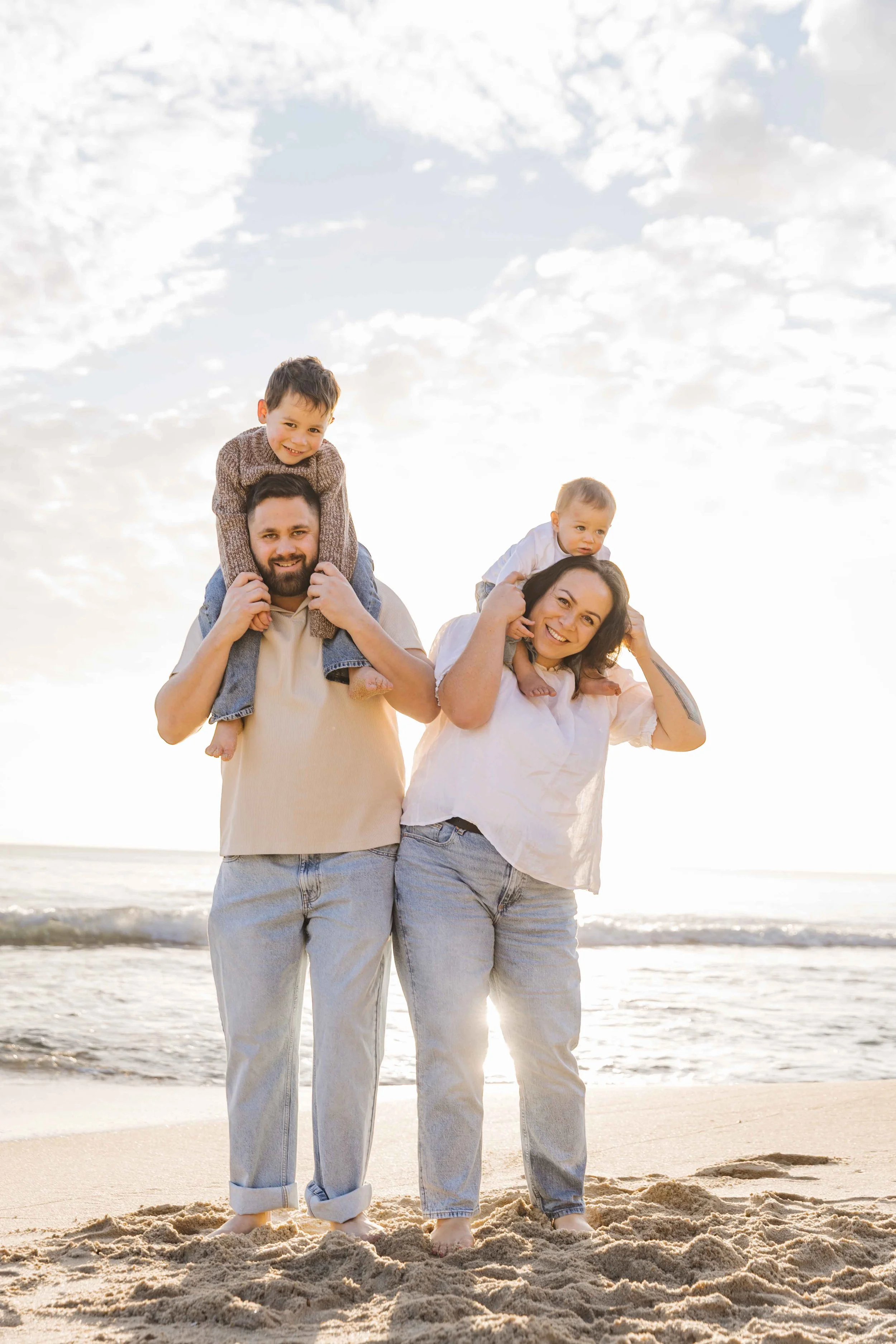 A family of four enjoying a day at the beach, with children riding on their parents' shoulders, all smiling, during sunset.