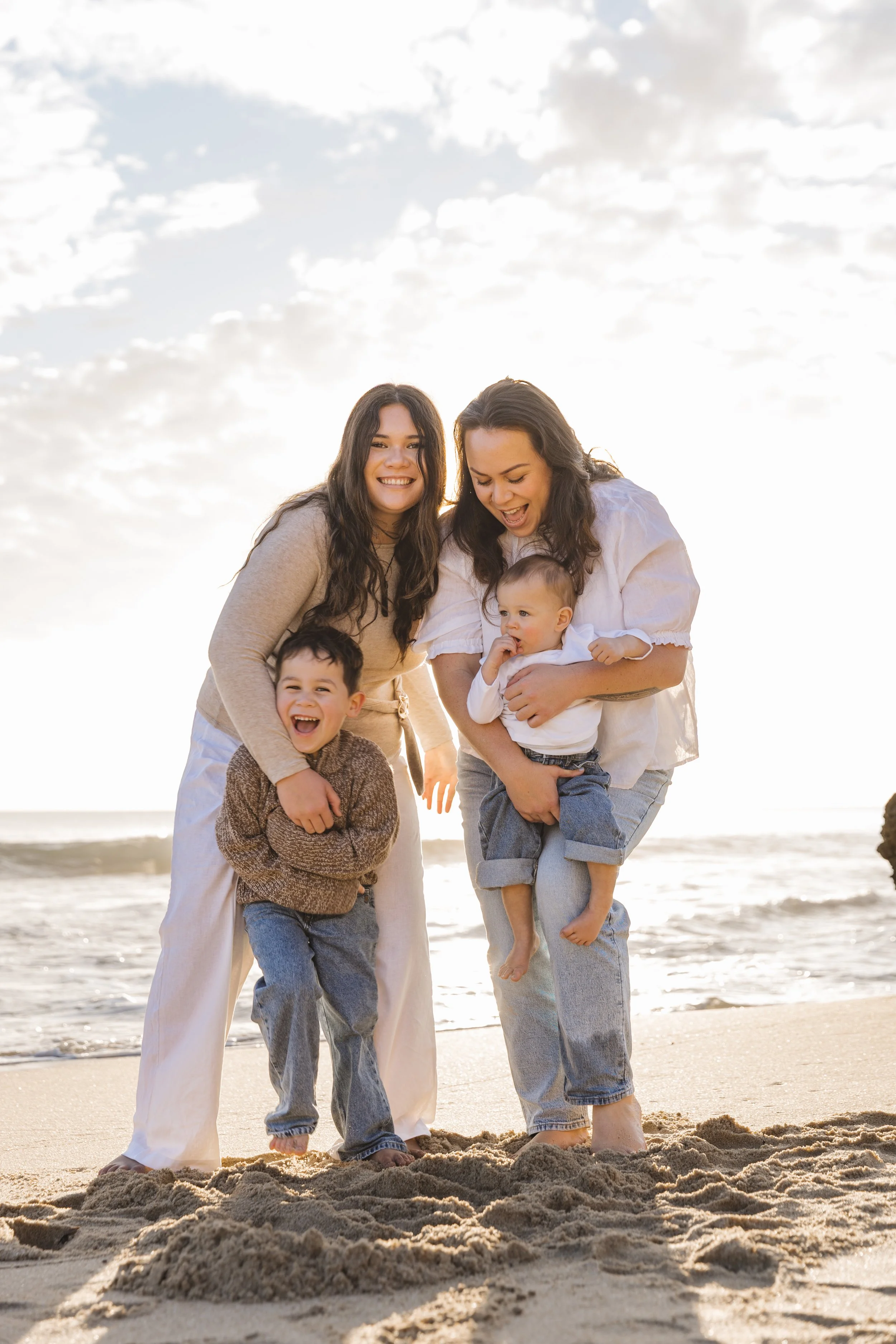 A group of four people, two women and two children, smiling and playing at the beach during sunset. The woman on the right is holding a young child, and the woman on the left is embracing a smiling boy. The sand and ocean are visible in the backgroun
