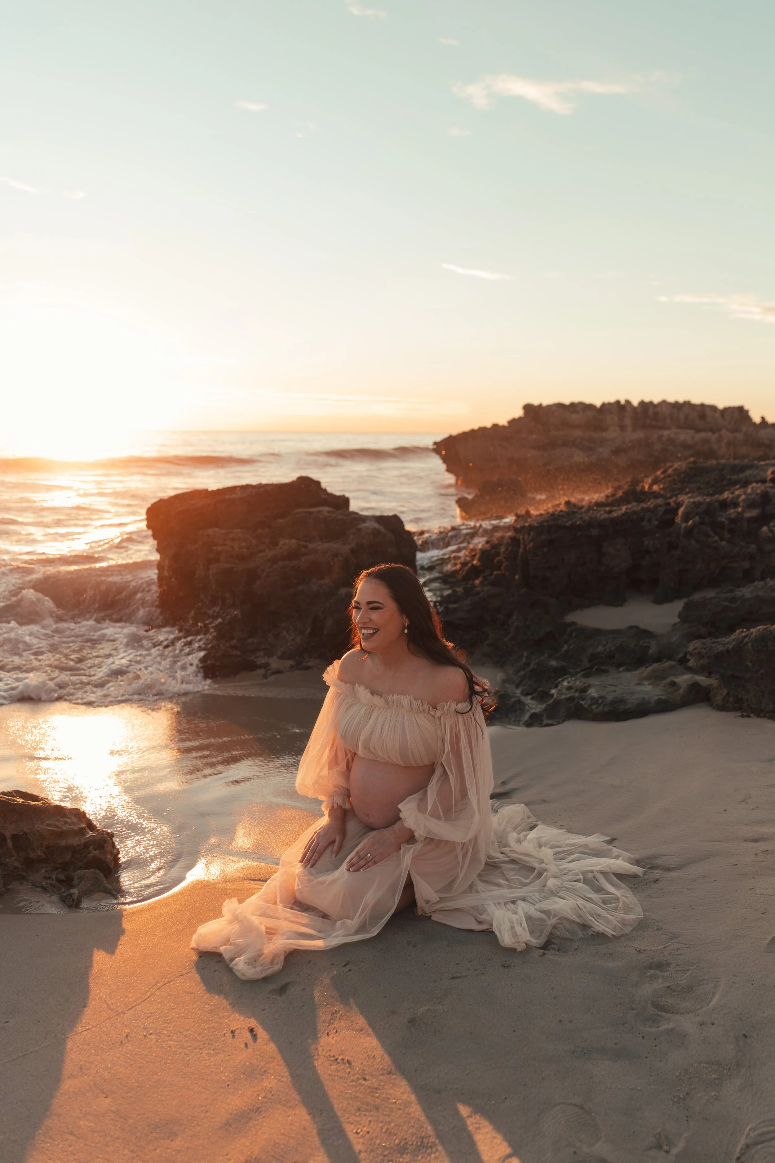 A pregnant woman smiling and kneeling on the sand at a beach during sunset, wearing a flowing, off-the-shoulder beige dress, with rocks and ocean waves in the background.