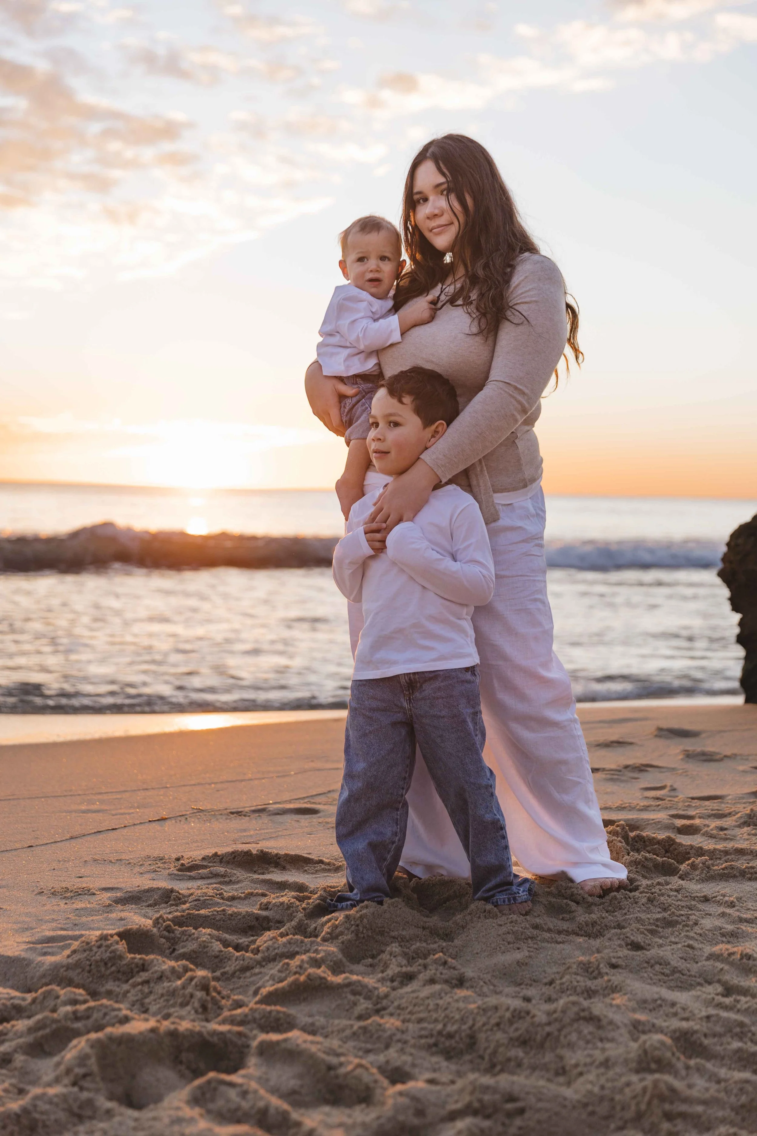 A woman with long dark hair standing on the beach at sunset, holding two young children, one in each arm, with ocean waves and rocks in the background.