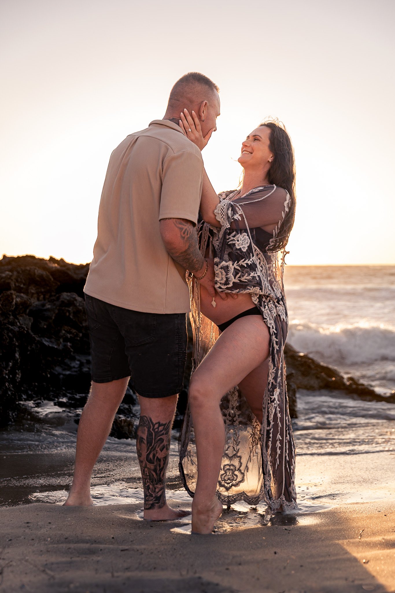 A man and woman standing close together on a beach at sunset, smiling and gazing into each other's eyes. The woman is pregnant, wearing a sheer, embroidered cover-up and black underwear, while the man has tattoos and is wearing a beige shirt and blac