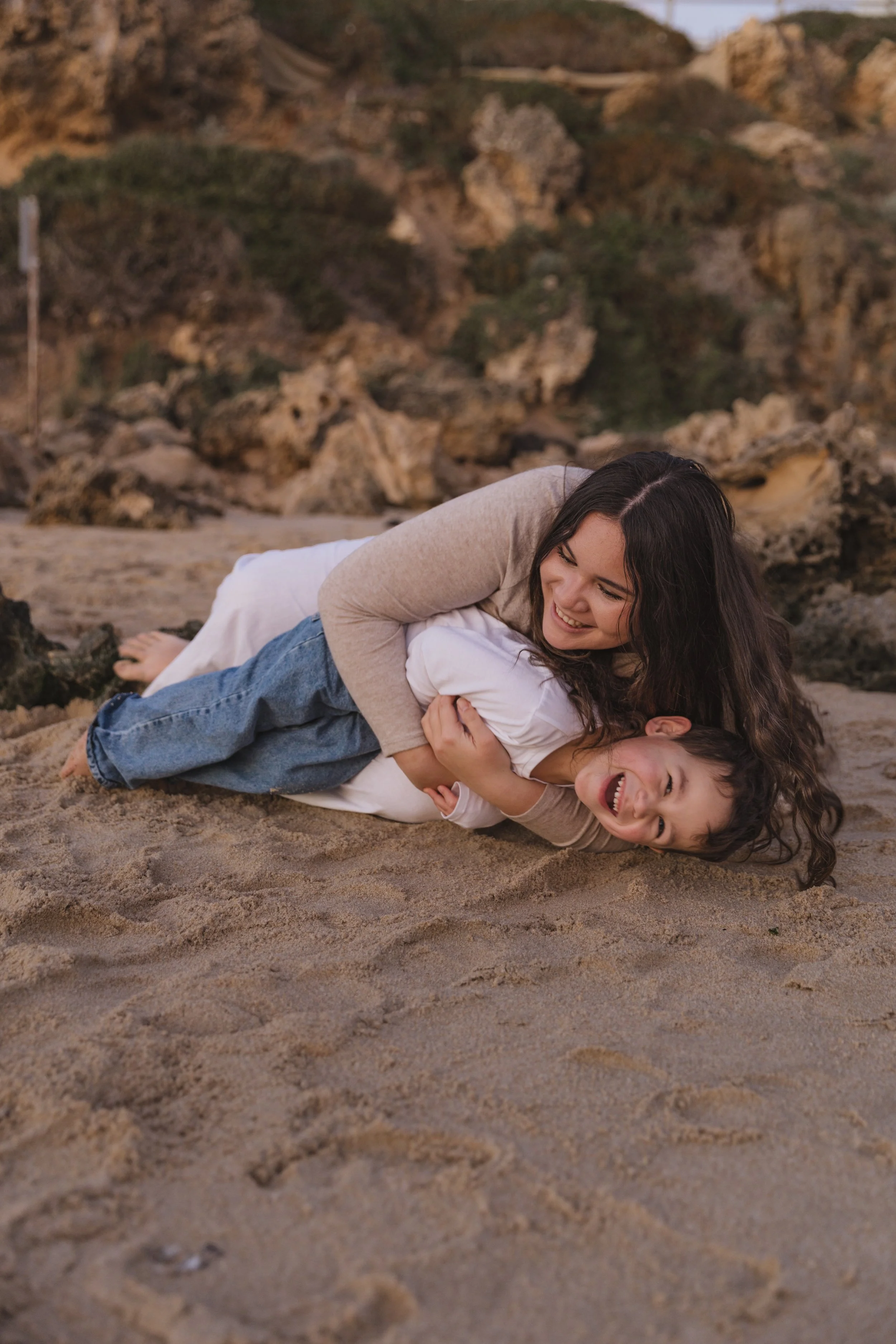 A sister and her younger brother playing and wrestling on the sandy beach, with rocky cliffs in the background.