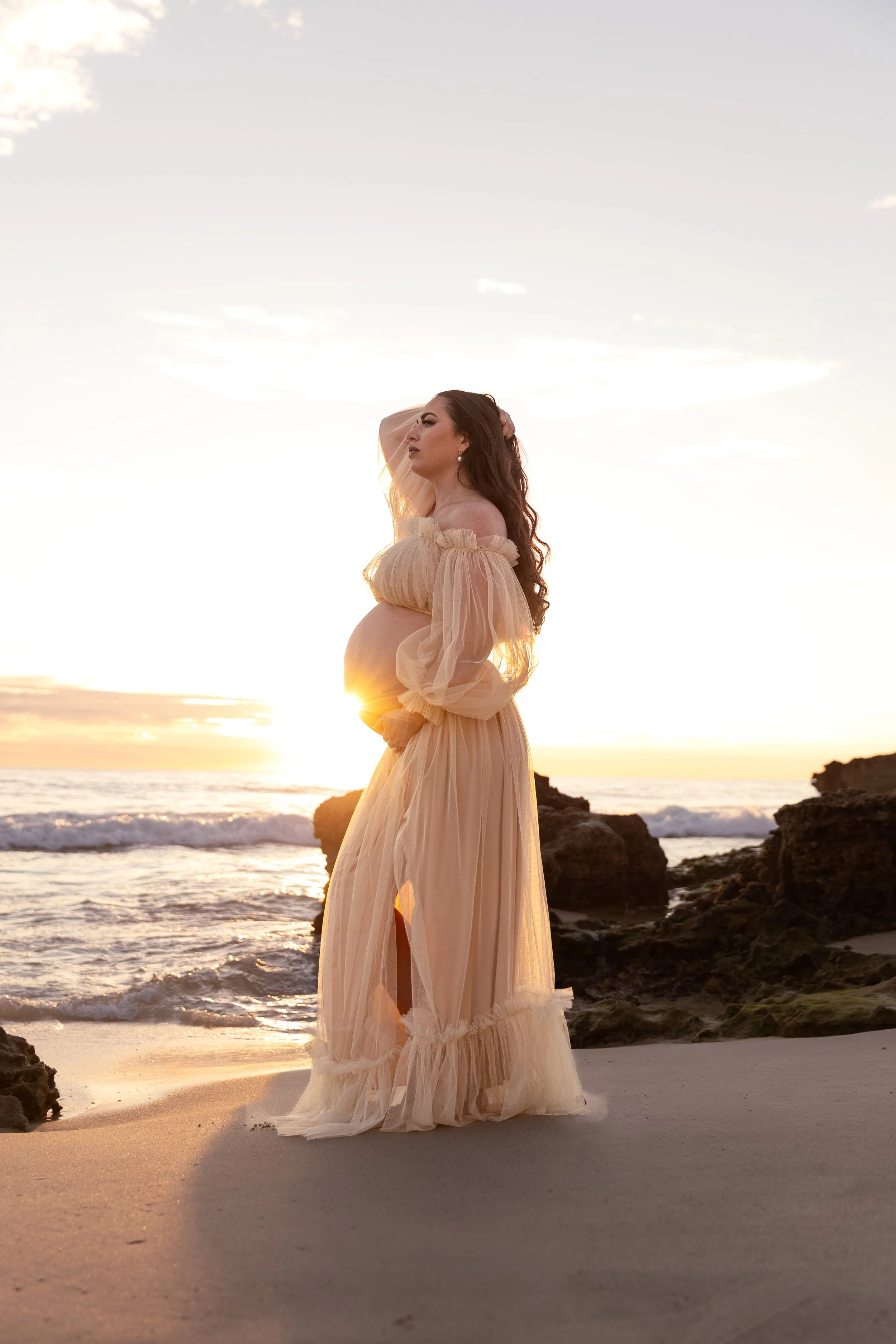 A woman standing on a beach at sunset, wearing a flowing beige dress with sheer puffed sleeves, with her hand on her head, and rocks in the background.