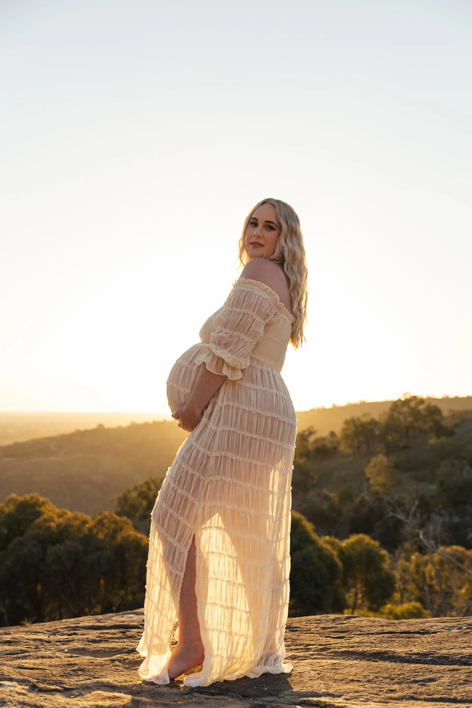 A woman in a flowing cream-colored dress standing barefoot on a rocky surface with a scenic sunset and landscape of trees and hills in the background.