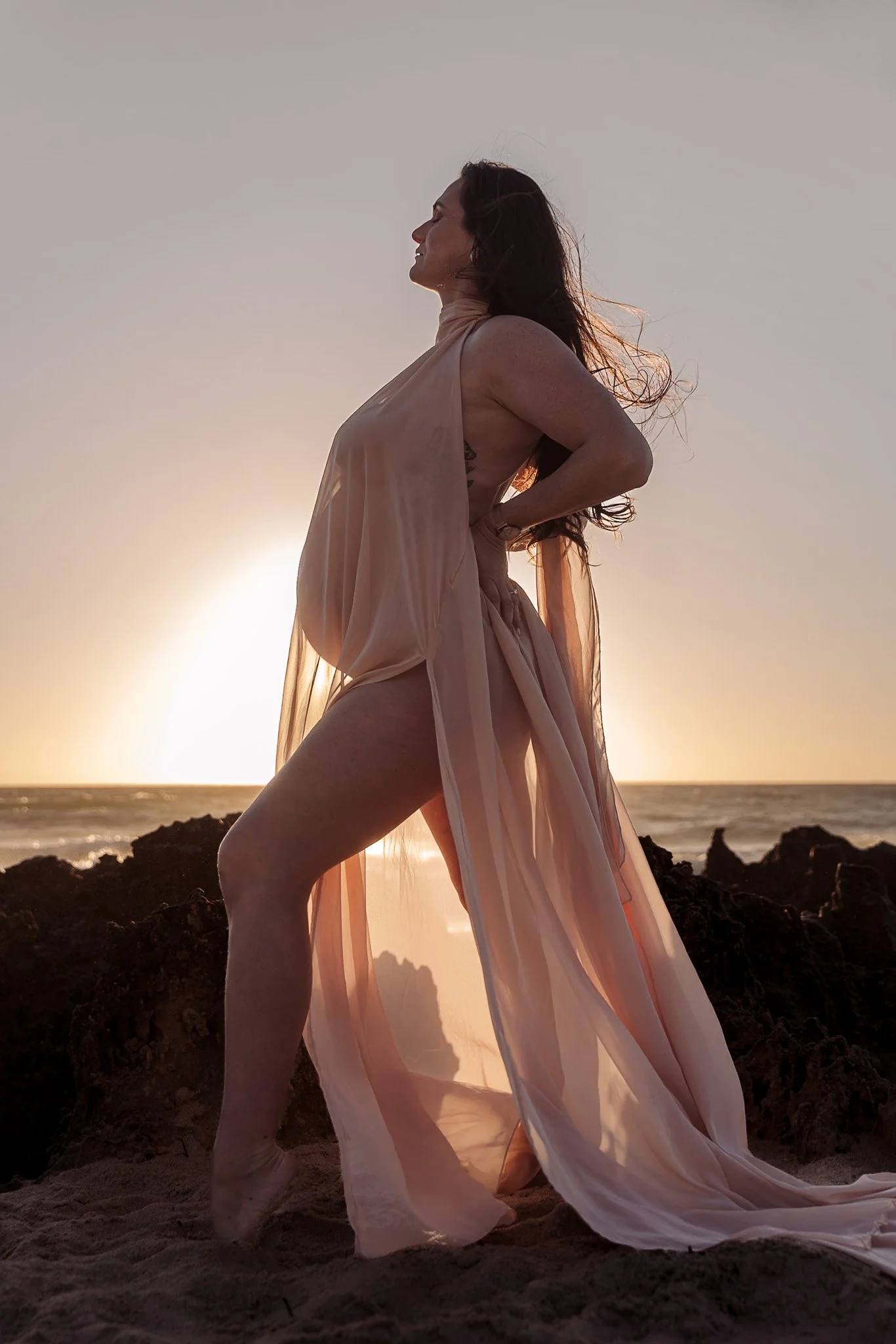 A woman in a flowing sheer pink dress stands on rocks at the beach with the sun setting behind her, creating a silhouette effect.